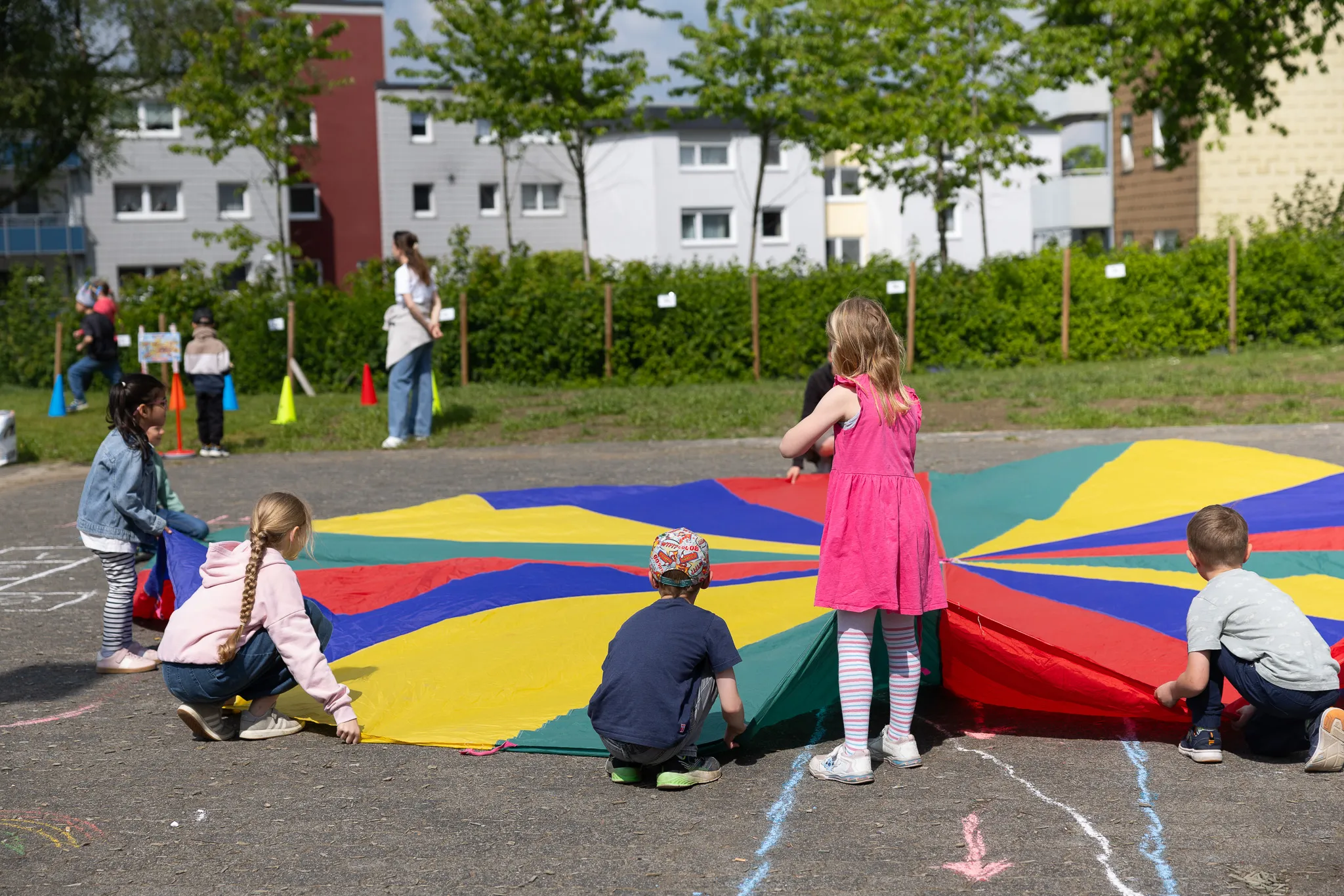 Kinder spielen auf einem bunten Fallschirm im Hof einer Schule, im Hintergrund ein Wohnhaus und Bäume.