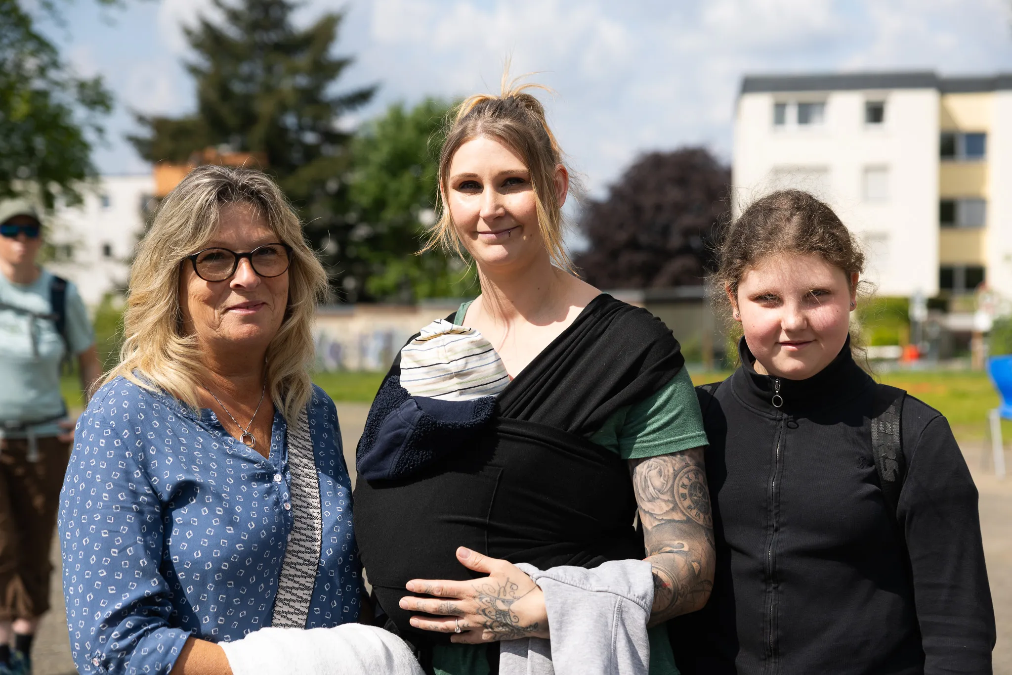Drei Frauen stehen lächelnd zusammen, eine davon trägt ein Baby in einem Tragetuch. Im Hintergrund sind weitere Menschen und Gebäude zu sehen.