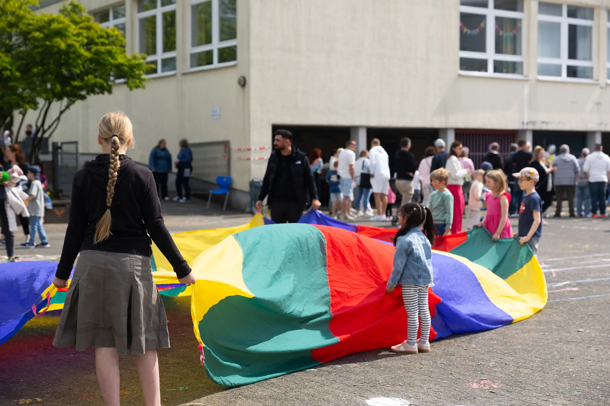 Kinder halten einen großen, farbenfrohen Fallschirm auf einem Schulhof, während im Hintergrund Menschen stehen und zusehen.