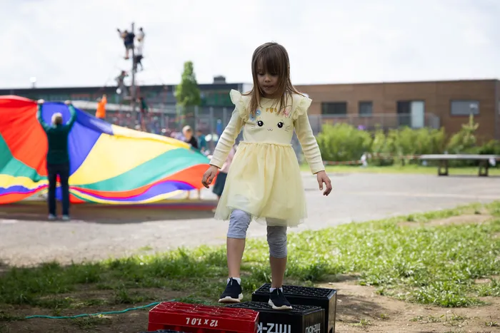 Ein junges Mädchen in gelbem Kleid balanciert auf einer schwarzen Kiste vor Regenbogenfallschirmen und einem Schulgebäude.