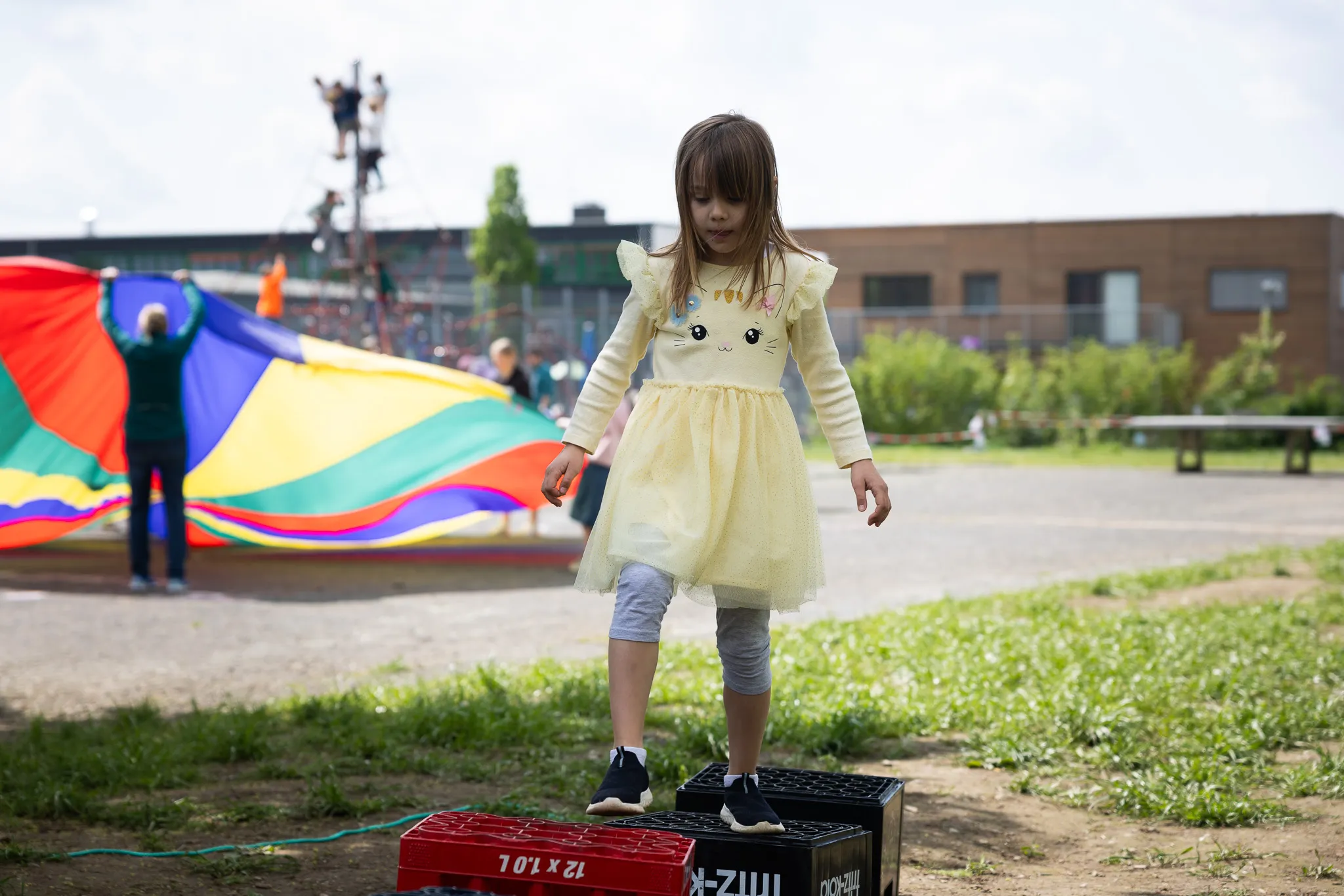 Ein junges Mädchen in gelbem Kleid balanciert auf einer schwarzen Kiste vor Regenbogenfallschirmen und einem Schulgebäude.