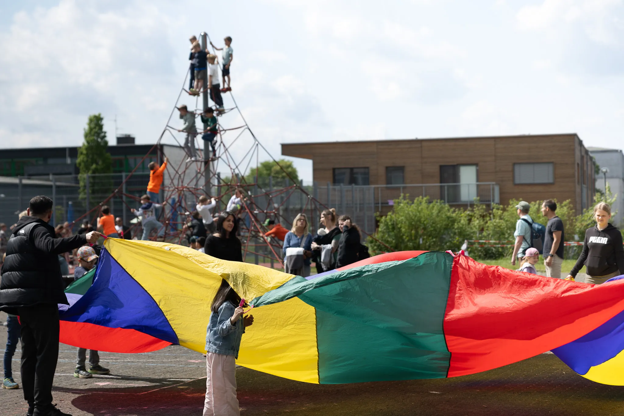 Kinder und Erwachsene halten einen großen Regenbogenfallschirm auf einem Schulhof, während andere Kinder auf einem Klettergerüst spielen. Im Hintergrund ein modernes Schulgebäude.