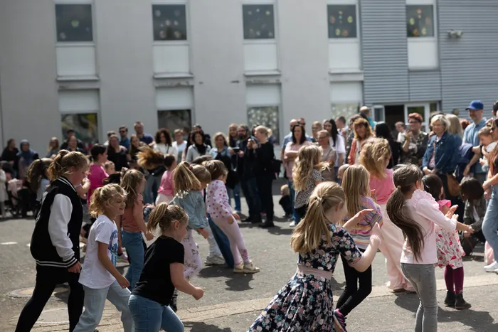 Eine Gruppe von Kindern tanzt im Freien vor einer Menschenmenge, die das Ereignis beobachtet. Im Hintergrund ein helles Gebäude mit mehreren Fenstern.