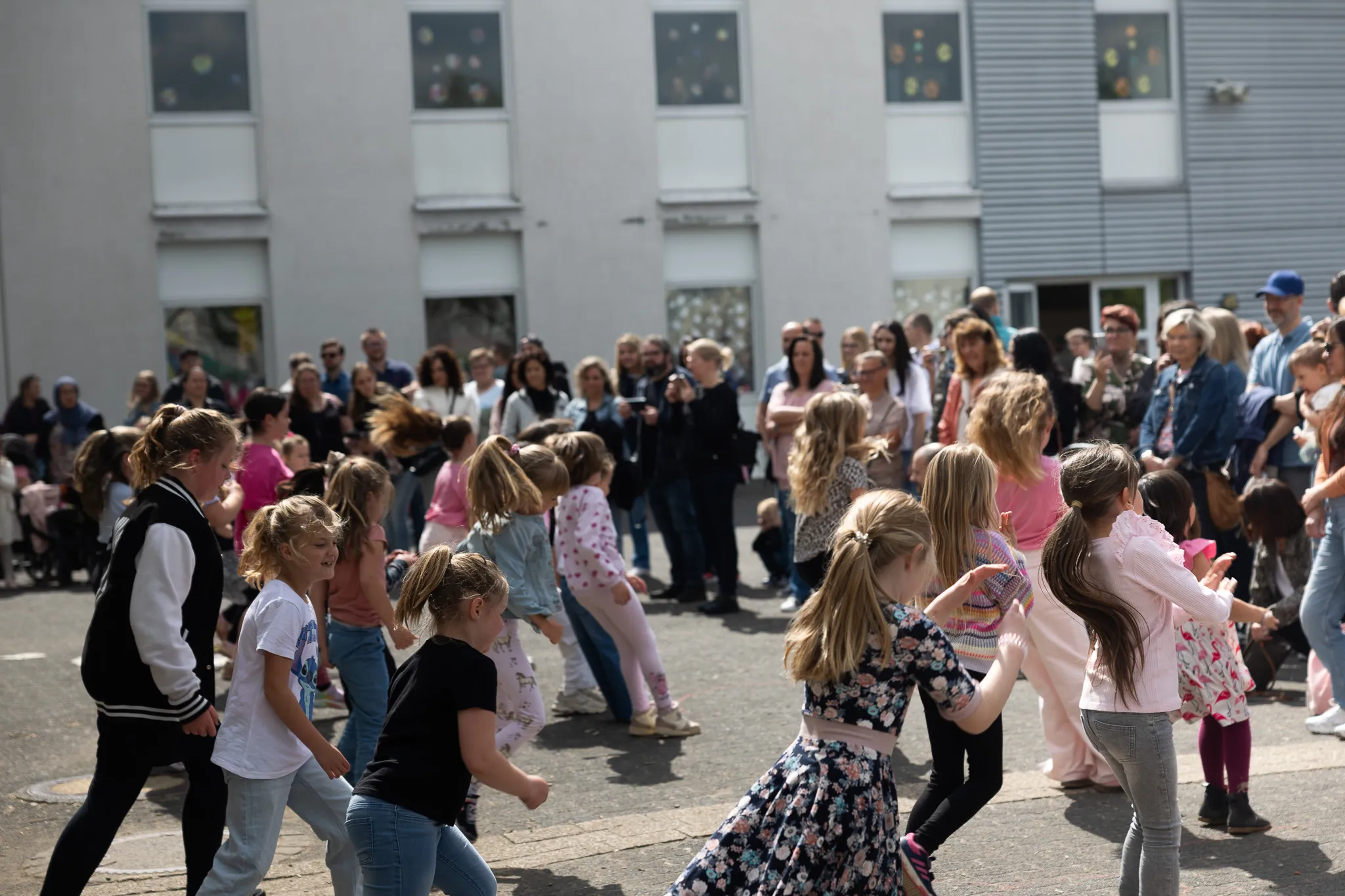 Eine Gruppe von Kindern tanzt im Freien vor einer Menschenmenge, die das Ereignis beobachtet. Im Hintergrund ein helles Gebäude mit mehreren Fenstern.
