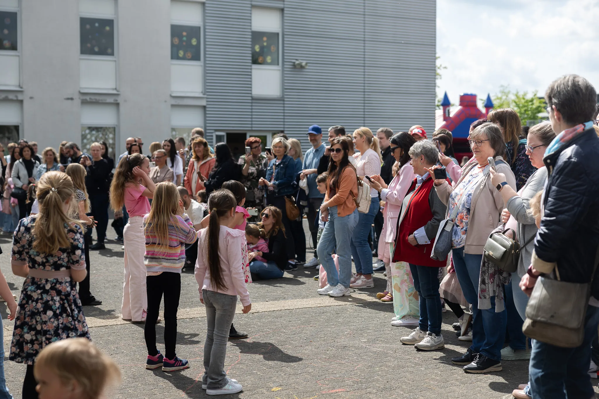 Eine Gruppe von Kindern und Erwachsenen steht auf einem Schulhof vor einem modernen Gebäude und beobachtet eine kleine Darbietung. Im Hintergrund sind weitere Menschen zu sehen, die das Fest besuchen.