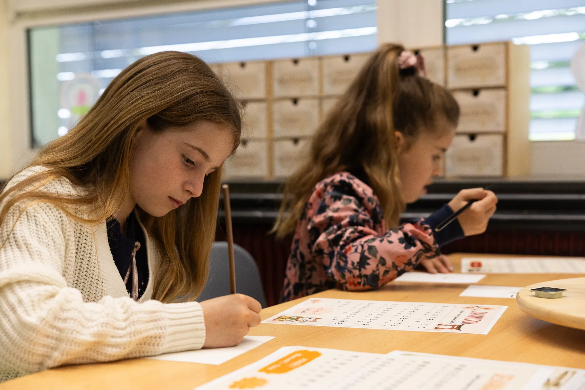 Zwei Schülerinnen sitzen an einem Tisch und schreiben konzentriert auf Arbeitsblättern während eines Schulfestes.