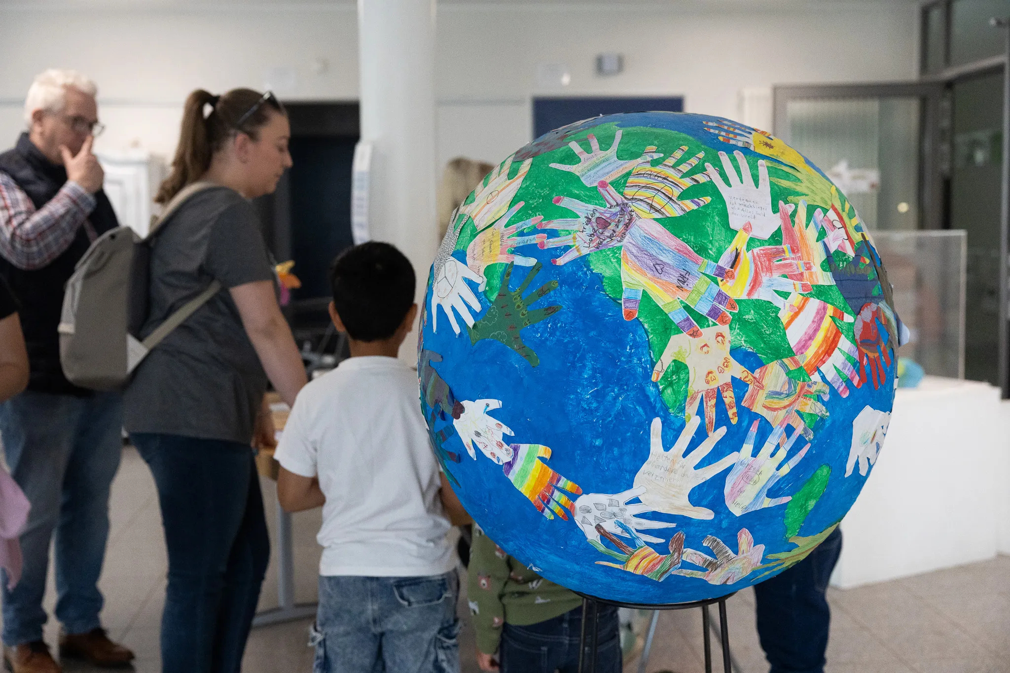 Eine große Weltkugel aus bemaltem Papier steht im Fokus, umgeben von Kindern und Erwachsenen, die sie betrachten. Die Kugel ist mit bunten Handabdrücken verziert.