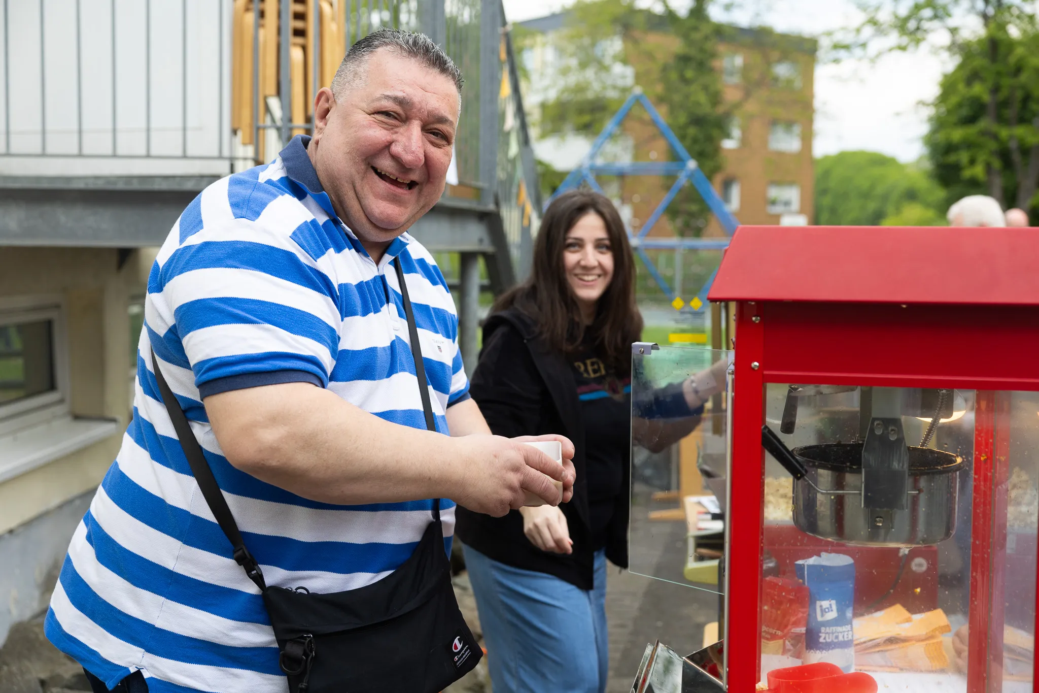 Ein Mann in gestreiftem Hemd hält eine Tasse vor einer Popcornmaschine, im Hintergrund eine lächelnde Frau und ein Spielplatz.