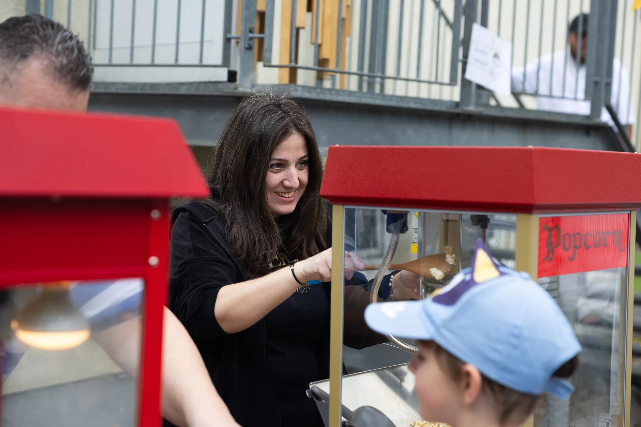 Frau bedient Popcornmaschine und reicht einem Kind mit blauer Kappe eine Tüte Popcorn.