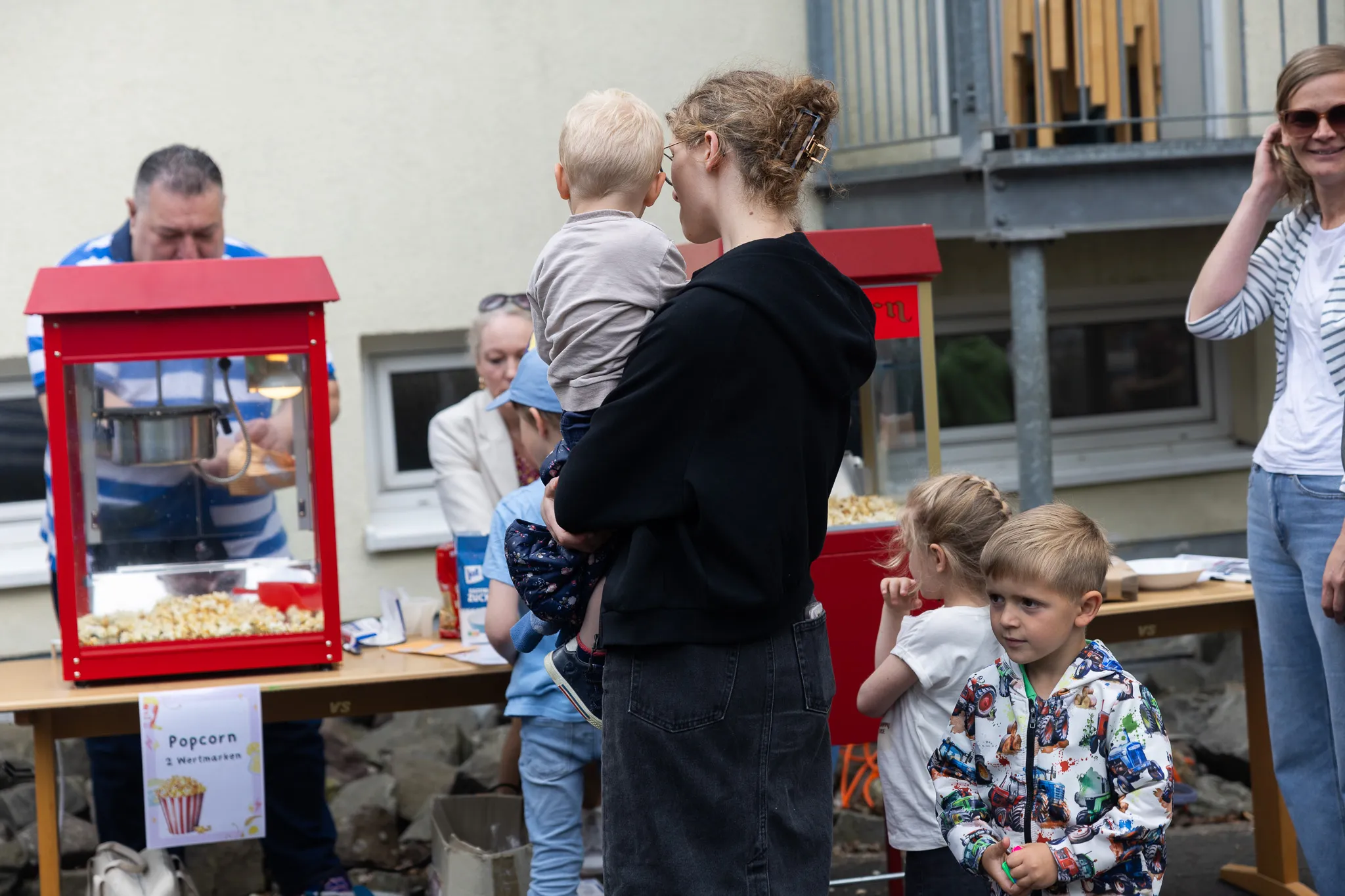 Eine Frau trägt ein blondes Kind auf dem Arm, während ein weiteres Kind vor ihr steht und zu einem Popcorn-Stand schaut.