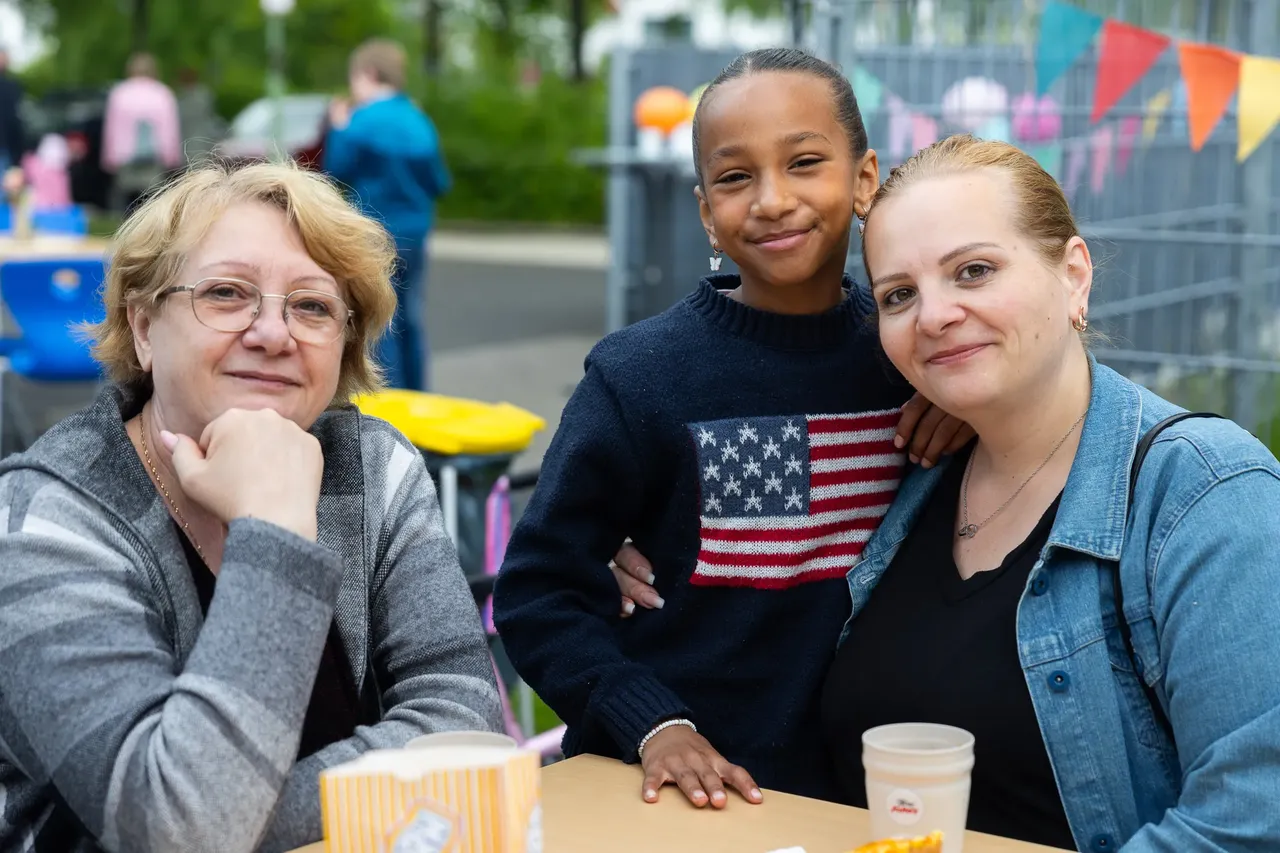 Drei Personen lächeln in die Kamera: eine ältere Frau links, ein Kind in der Mitte mit einem Pullover mit amerikanischer Flagge und eine jüngere Frau rechts. Im Hintergrund sind weitere Menschen und bunte Flaggen zu sehen.