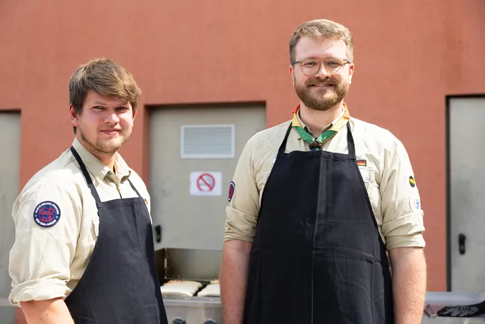 Zwei Männer mit Schürzen stehen vor einem Grillgerät. Im Hintergrund ist ein roter Gebäudeteil mit einem Lüftungsgitter und einem Verbotsschild zu sehen.
