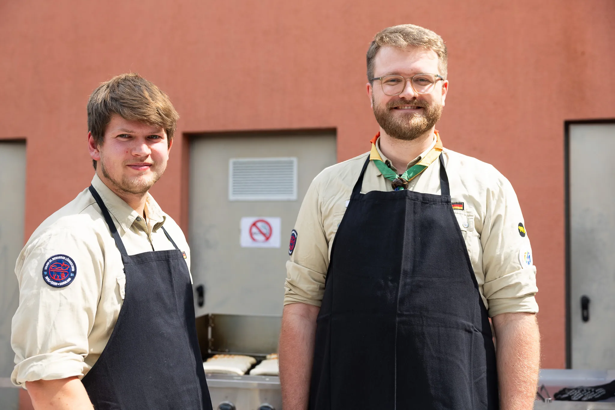 Zwei Männer mit Schürzen stehen vor einem Grillgerät. Im Hintergrund ist ein roter Gebäudeteil mit einem Lüftungsgitter und einem Verbotsschild zu sehen.