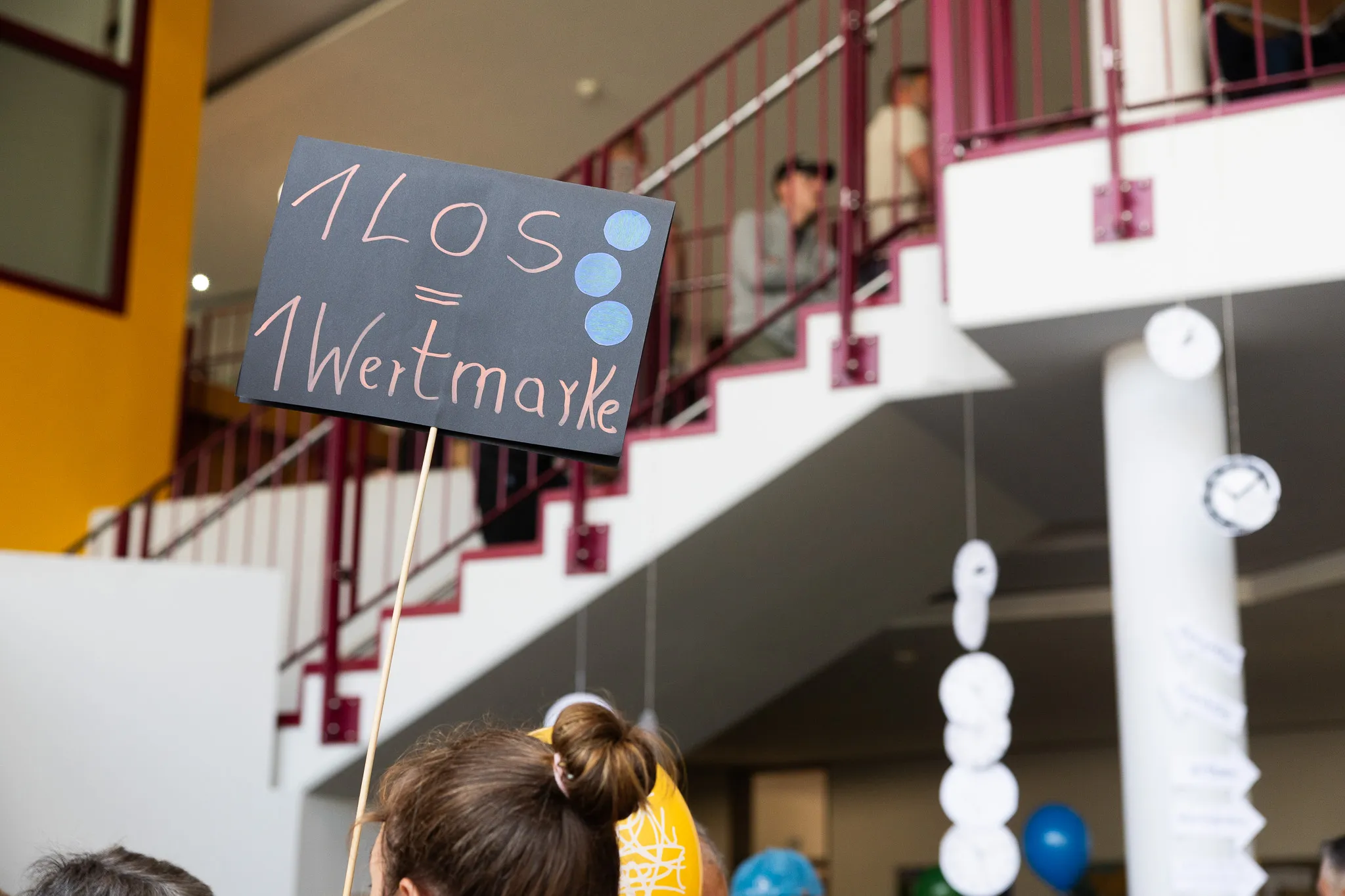 Schwarzes Schild mit der Aufschrift „1LOS = 1 Wertmarke“ wird auf einer Treppe in einem hellen Schulgebäude gehalten.