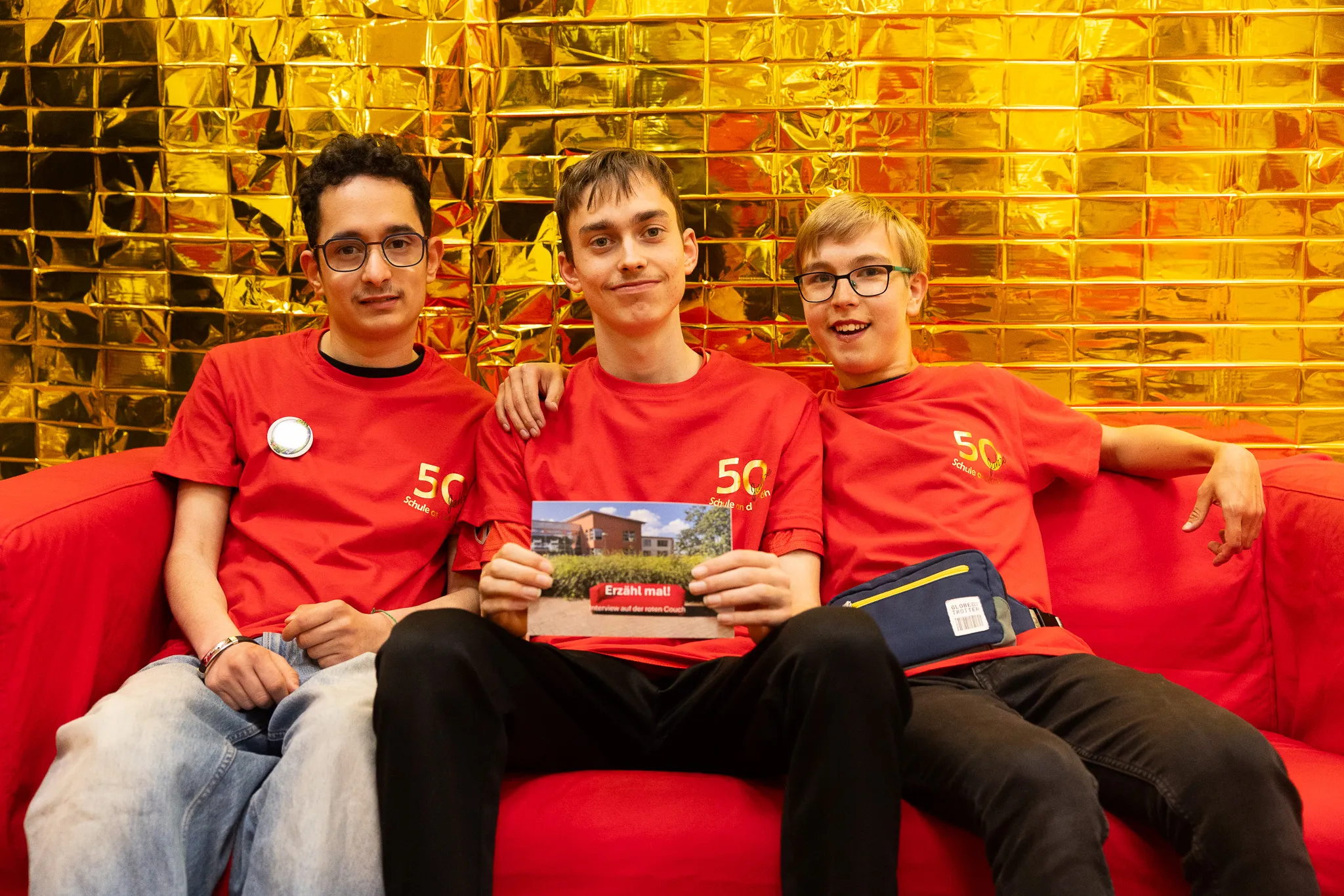 Drei junge Menschen sitzen lächelnd auf einem roten Sofa und halten ein Buch mit einem Bild des Schulgebäudes hoch. Sie tragen rote T-Shirts mit der Aufschrift „50 Schule“.