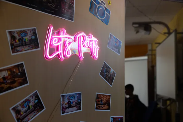 Leuchtende Neonreklame mit der Aufschrift „Let’s Party“ an einer Wand mit Fotos von Schülern und Veranstaltungen.