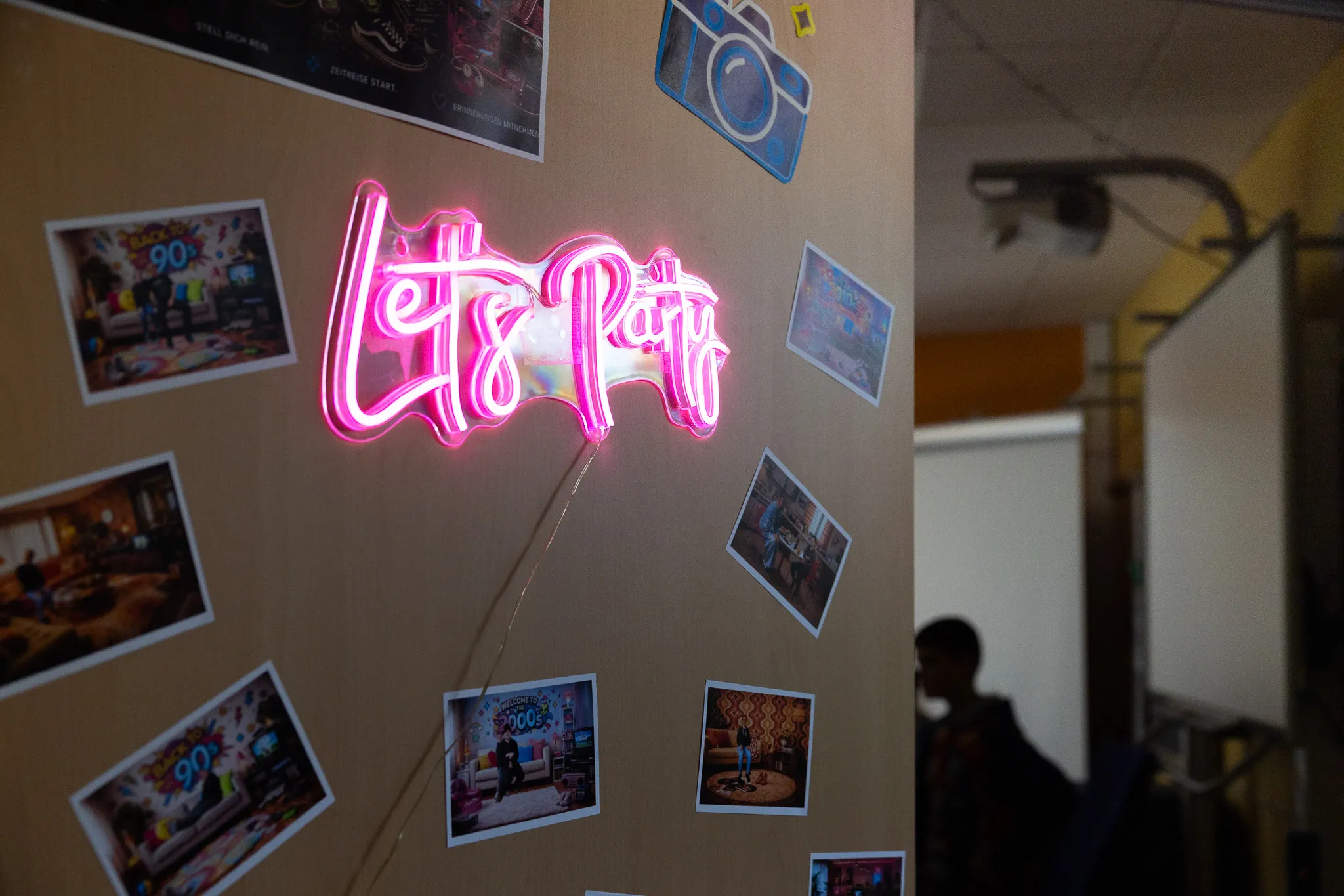 Leuchtende Neonreklame mit der Aufschrift „Let’s Party“ an einer Wand mit Fotos von Schülern und Veranstaltungen.