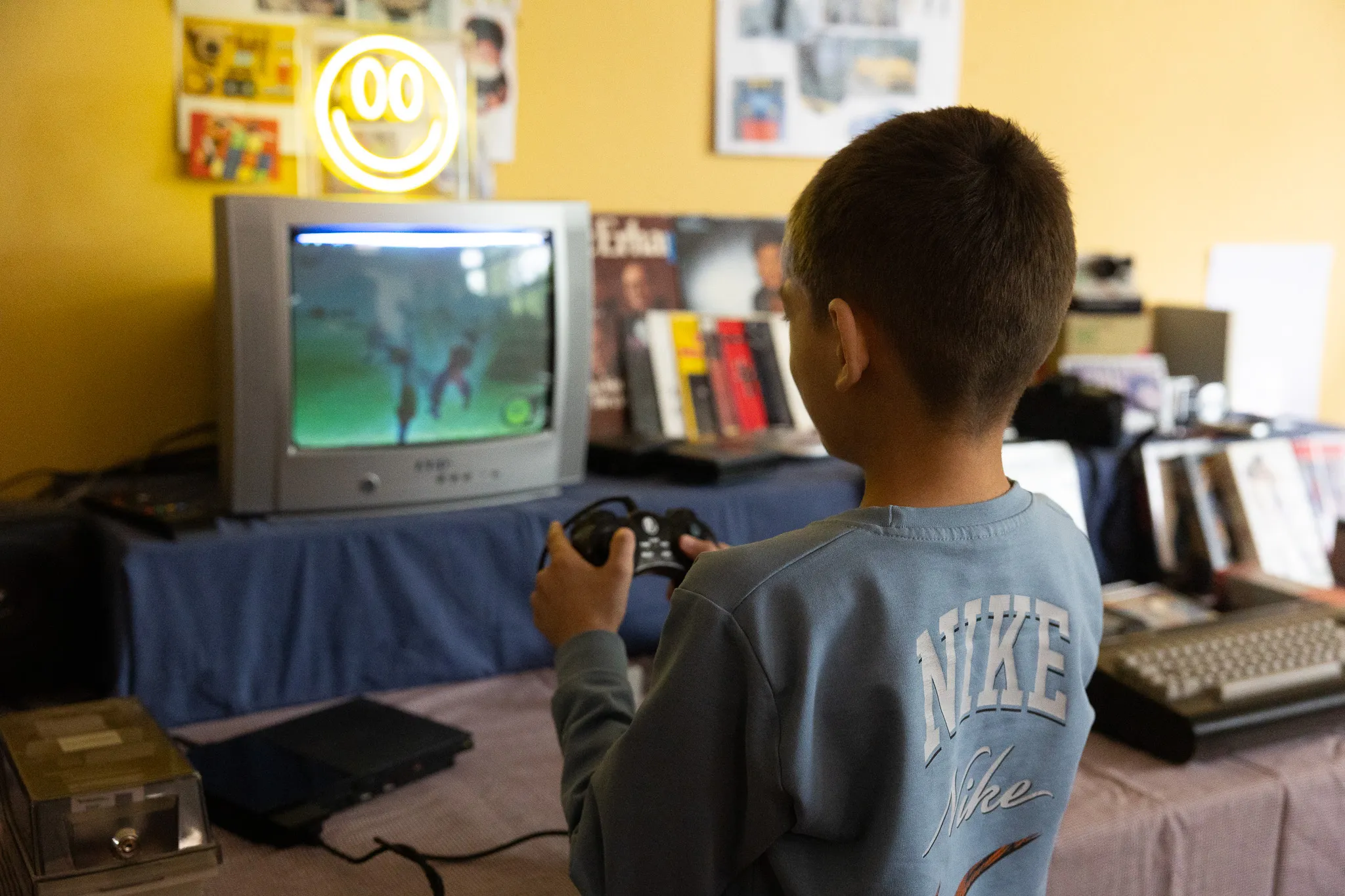 Junge spielt Videospiel auf einer älteren Konsole, im Hintergrund Bücher und ein Neon-Lächeln.