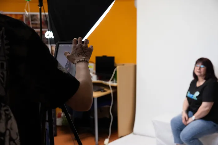 Eine Frau sitzt vor einer weißen Wand und wird von einer Person mit einem Tablet gefilmt. Im Hintergrund sind Studiobeleuchtung und Büromöbel zu sehen.