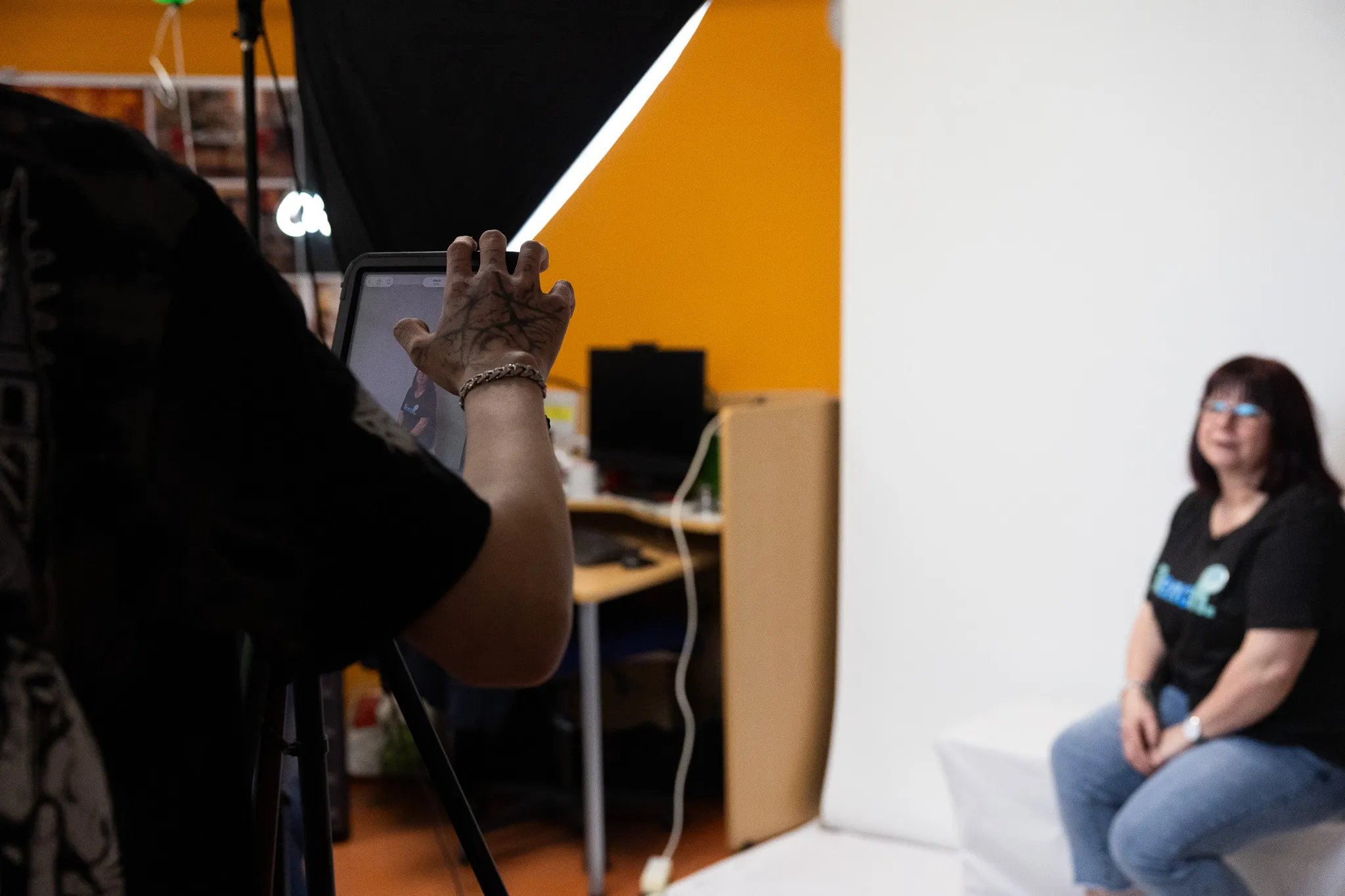 Eine Frau sitzt vor einer weißen Wand und wird von einer Person mit einem Tablet gefilmt. Im Hintergrund sind Studiobeleuchtung und Büromöbel zu sehen.