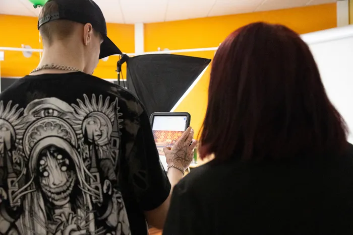 Eine Person mit rotem Haar fotografiert mit einem Tablet ein Kunstwerk an der Wand, während eine andere Person mit Kappe und aufwendigem T-Shirt im Hintergrund steht.