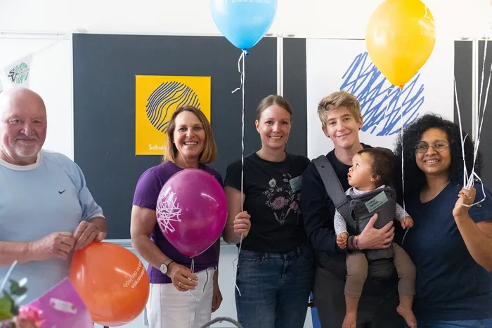 Fünf Personen stehen lächelnd mit bunten Luftballons vor einer Wand mit dem Schriftzug „Schule an der Höh“ und einem abstrakten, farbigen Muster.