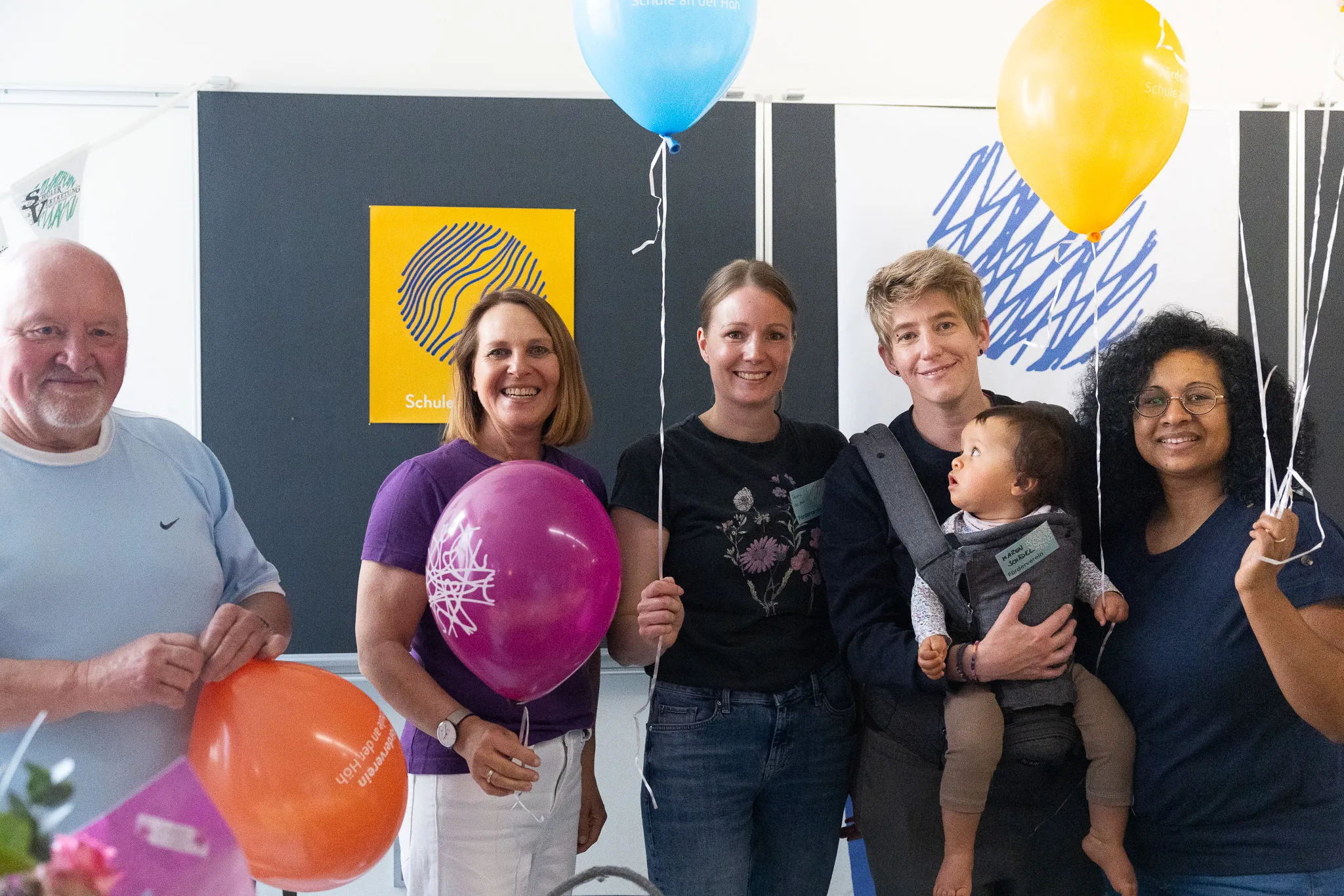 Fünf Personen stehen lächelnd mit bunten Luftballons vor einer Wand mit dem Schriftzug „Schule an der Höh“ und einem abstrakten, farbigen Muster.