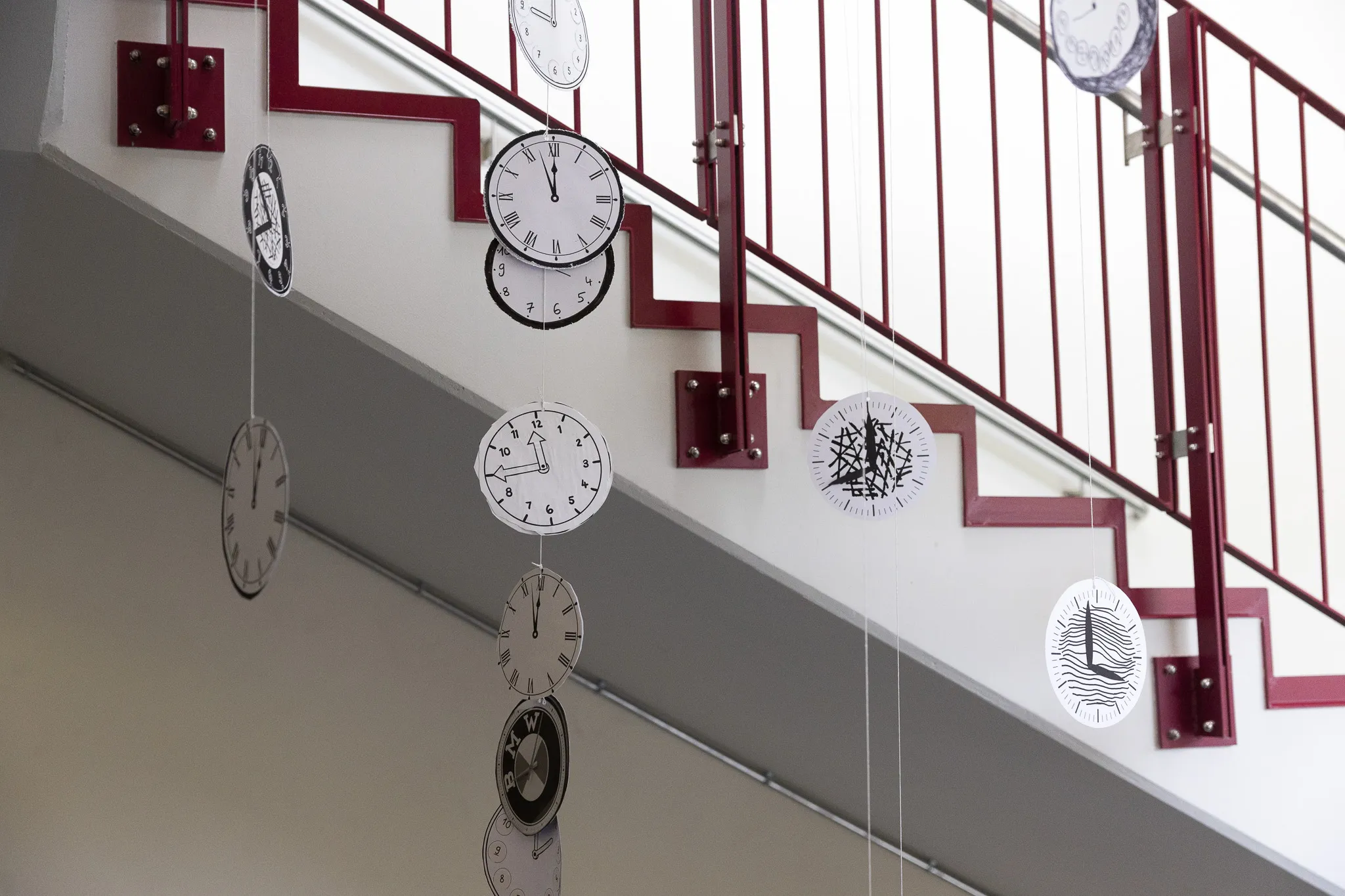Mehrere stilisierte Uhren hängen an dünnen Fäden von der Decke einer hellen Treppe mit rotem Geländer.