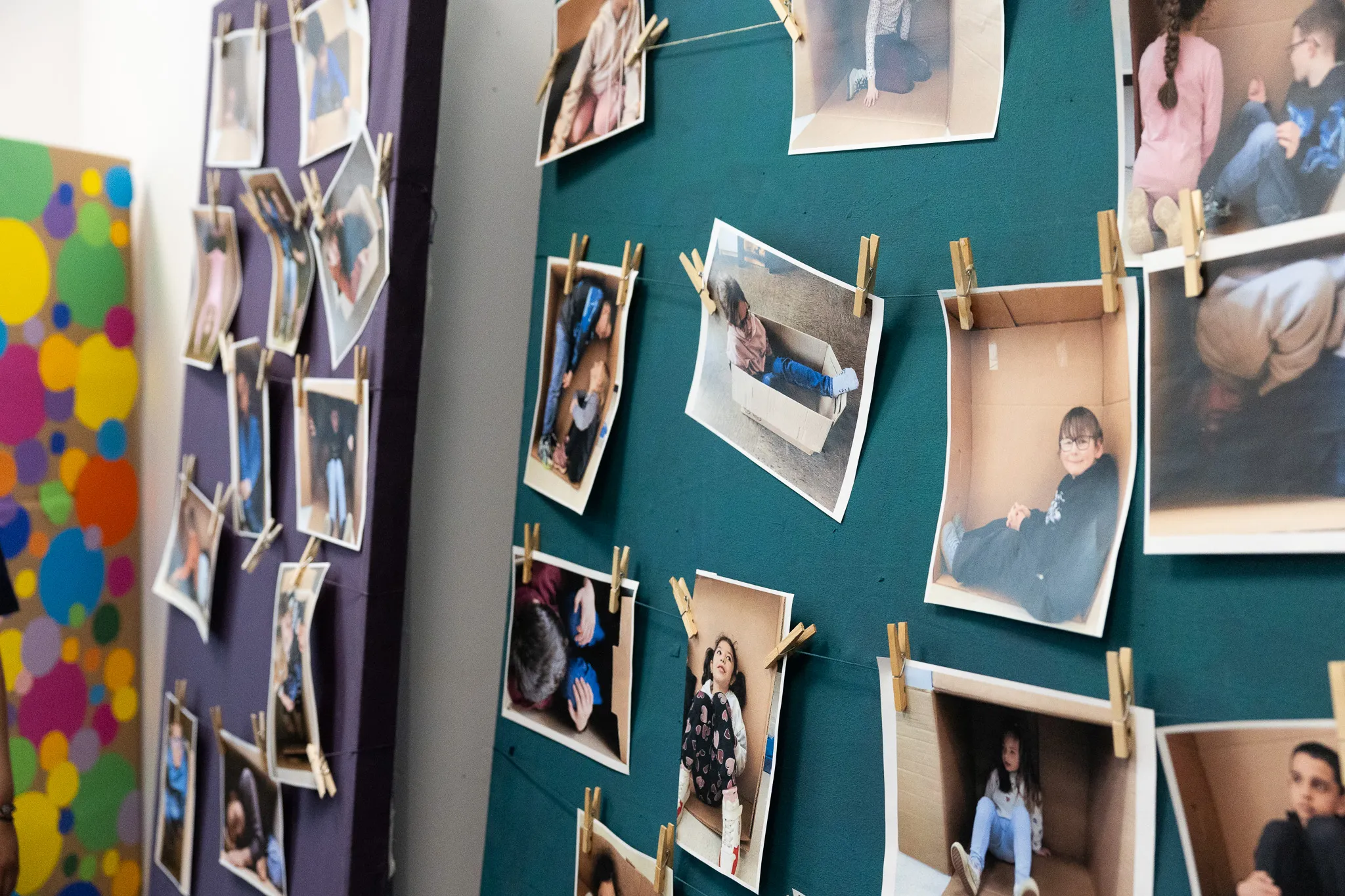 An einer Pinnwand hängen zahlreiche Kinderfotos, die mit Wäscheklammern befestigt sind. Die Fotos zeigen Kinder in verschiedenen Posen, darunter auch einige, die in Kartons sitzen.