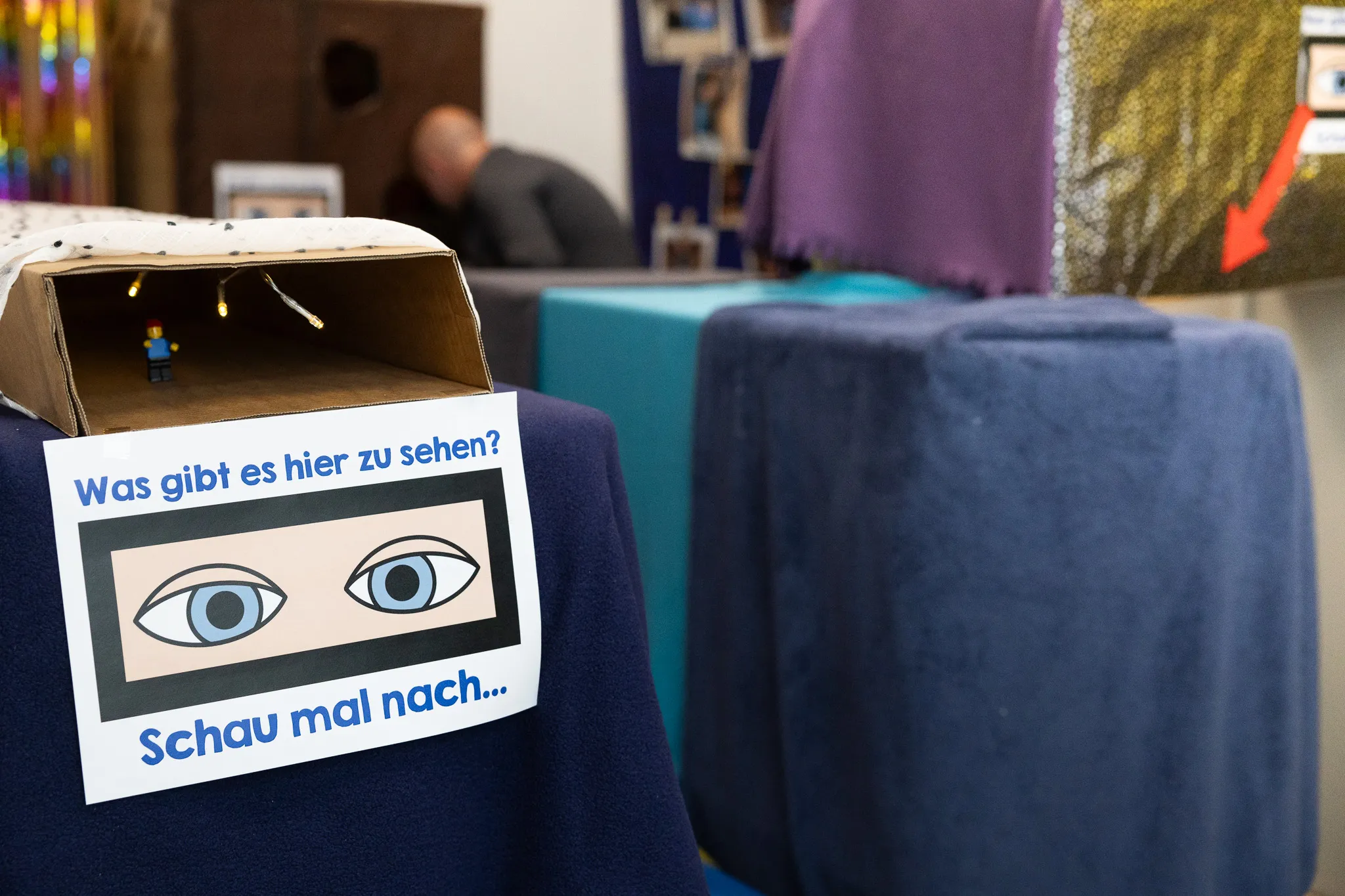 Ein selbstgebauter Karton mit zwei Augen und der Aufschrift „Was gibt es hier zu sehen? Schau mal nach…“ steht auf einem blauen Tisch.