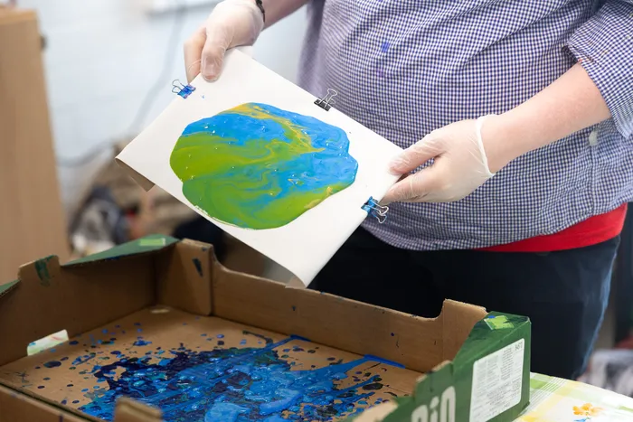 Hände mit weißen Handschuhen halten ein farbiges Bild der Erde mit blauen, grünen und gelben Farbtönen, befestigt mit Büroklammern. Darunter befindet sich eine Schachtel mit Farbresten.