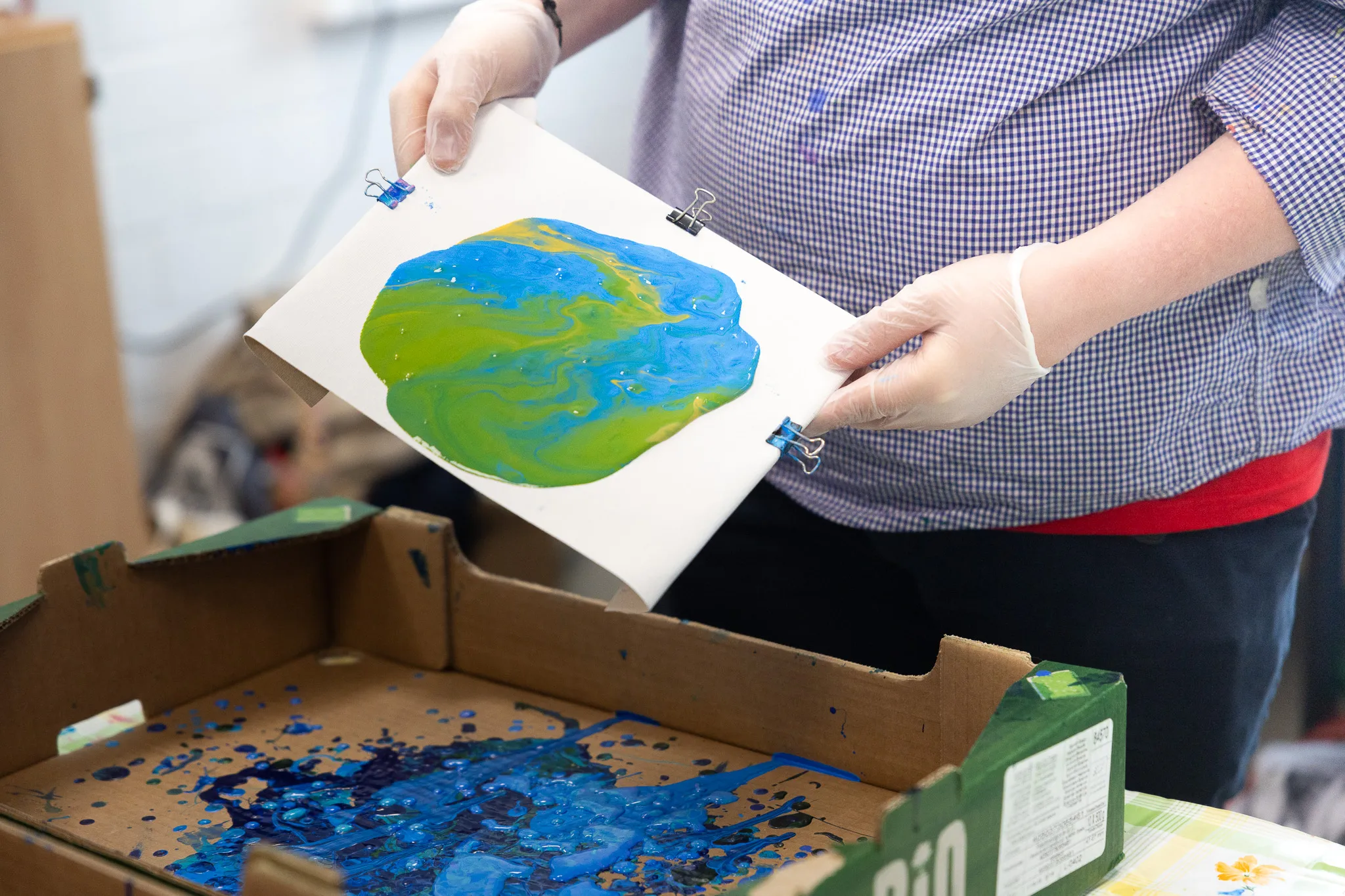 Hände mit weißen Handschuhen halten ein farbiges Bild der Erde mit blauen, grünen und gelben Farbtönen, befestigt mit Büroklammern. Darunter befindet sich eine Schachtel mit Farbresten.