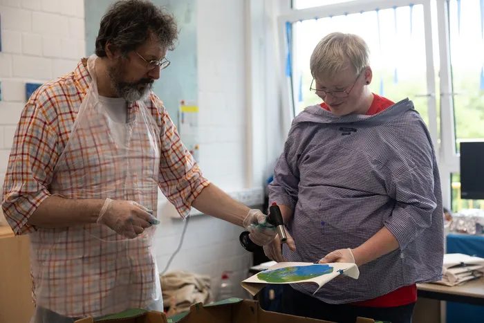 Zwei Personen arbeiten gemeinsam an einem farbigen Bild, möglicherweise im Rahmen eines Kunstprojekts zur Schulfeier.