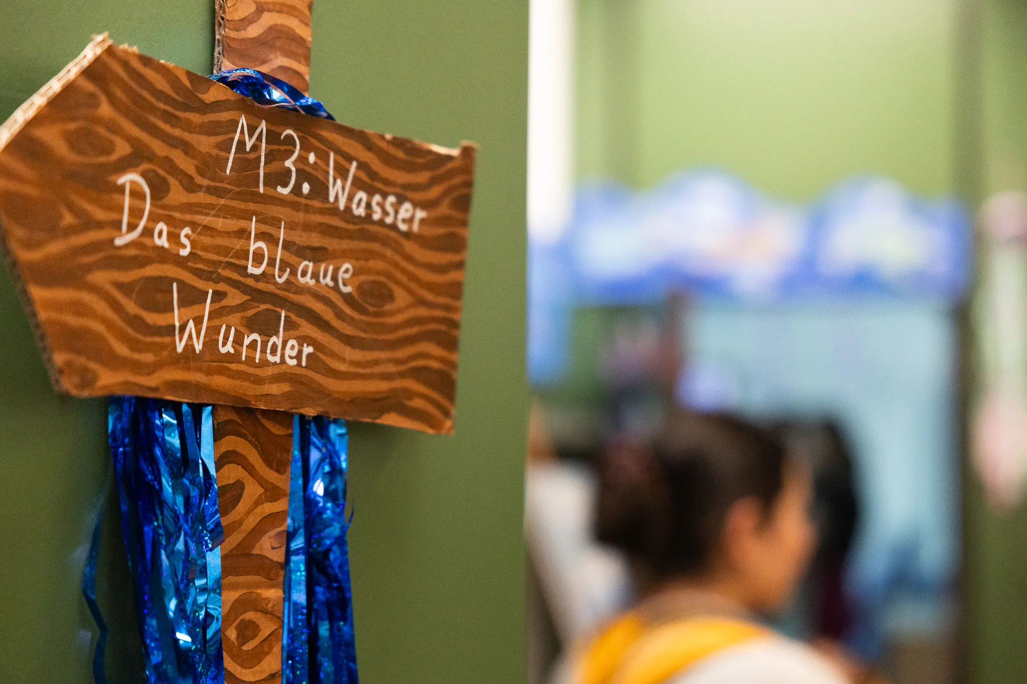 Holzschild mit der Aufschrift „M3: Wasser – Das blaue Wunder“ und blauen Lamellen, im Hintergrund verschwommene Personen in einer Schulaula.