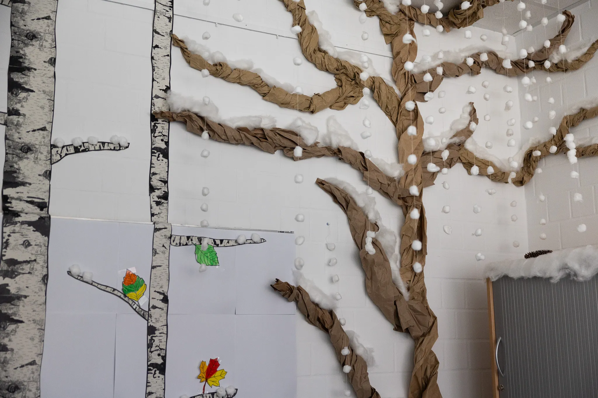 Wanddekoration mit Birkenstämmen und Ästen aus Papier, an denen Blätter und weiße Kugeln befestigt sind.