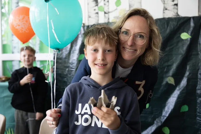 Frau und Junge lächeln in die Kamera, halten Luftballons fest. Im Hintergrund ein weiteres Kind und eine dekorierte Wand.
