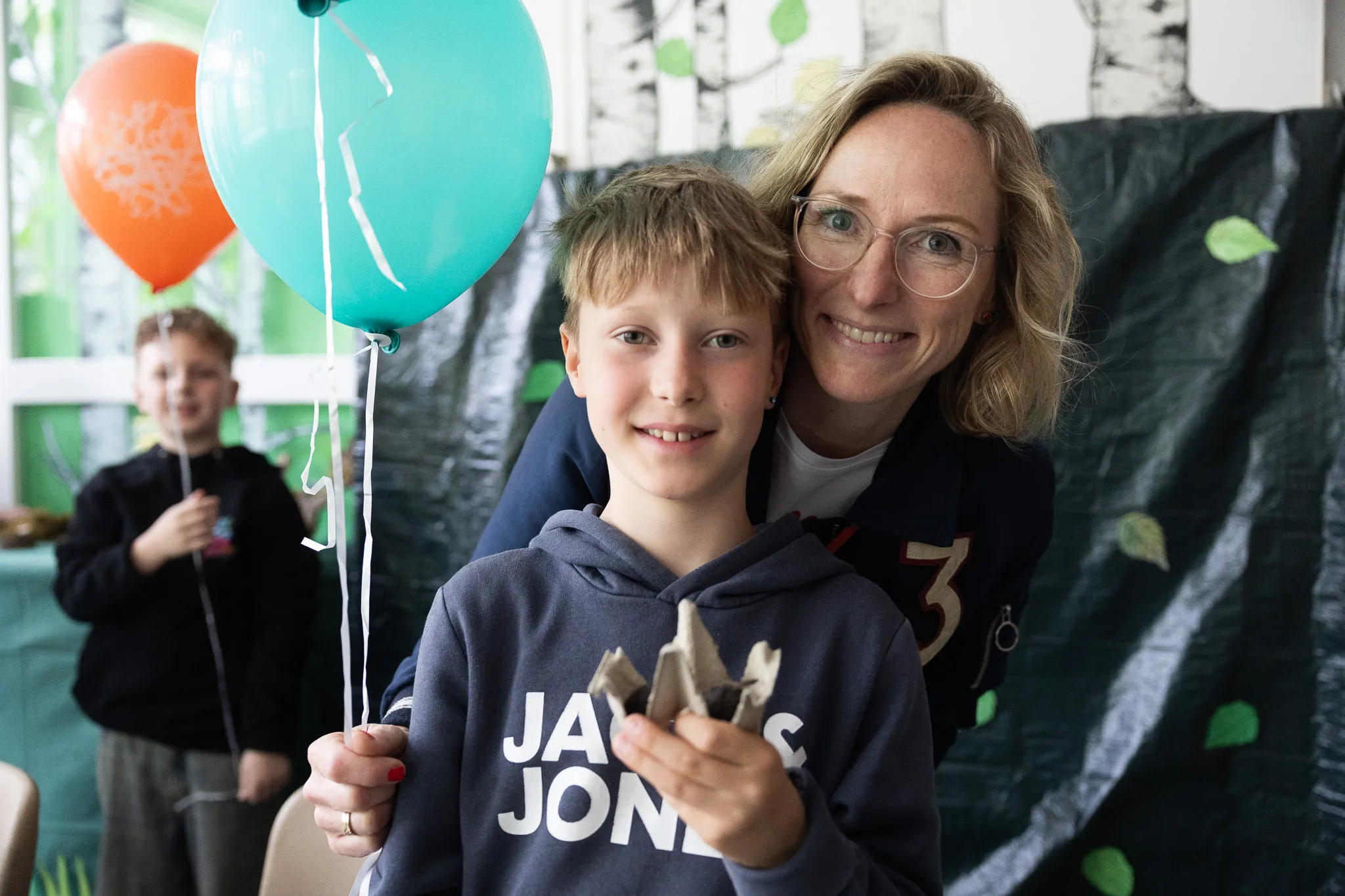 Frau und Junge lächeln in die Kamera, halten Luftballons fest. Im Hintergrund ein weiteres Kind und eine dekorierte Wand.