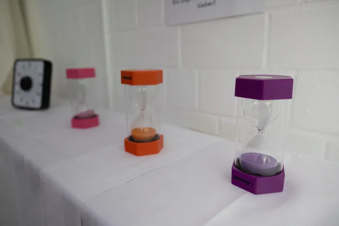 Drei Sanduhren in Pink, Orange und Lila stehen auf einem weißen Tisch vor einer hellen Wand mit Kacheln.