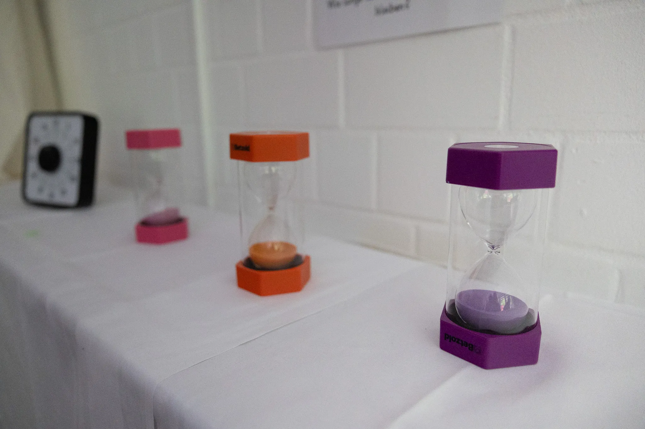 Drei Sanduhren in Pink, Orange und Lila stehen auf einem weißen Tisch vor einer hellen Wand mit Kacheln.