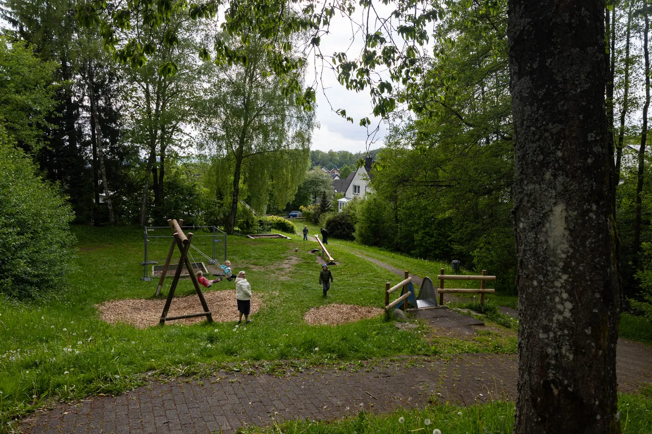 Spielplatz mit Schaukeln und Klettergerüsten, umgeben von Bäumen und Grünflächen. Im Hintergrund sind Häuser zu sehen.