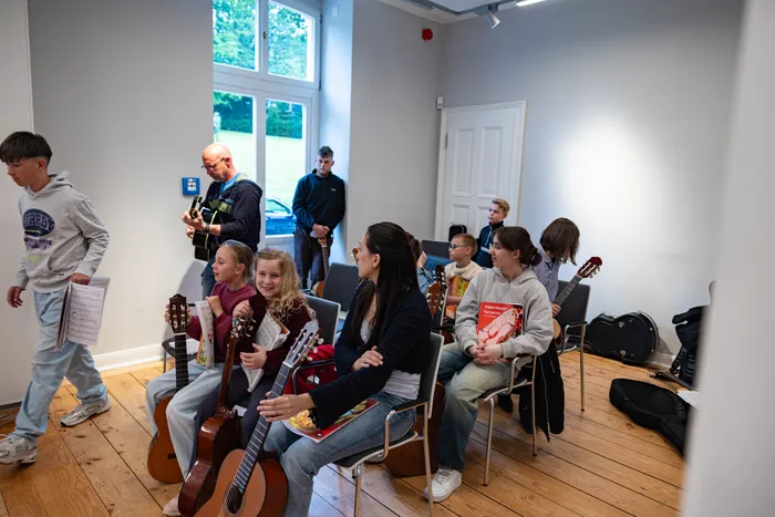 Mehrere Kinder und Jugendliche sitzen oder stehen in einem hellen Raum mit Gitarren und Notenblättern, während eine Frau eine Gitarre spielt und ein Mann im Hintergrund ebenfalls eine Gitarre hält.