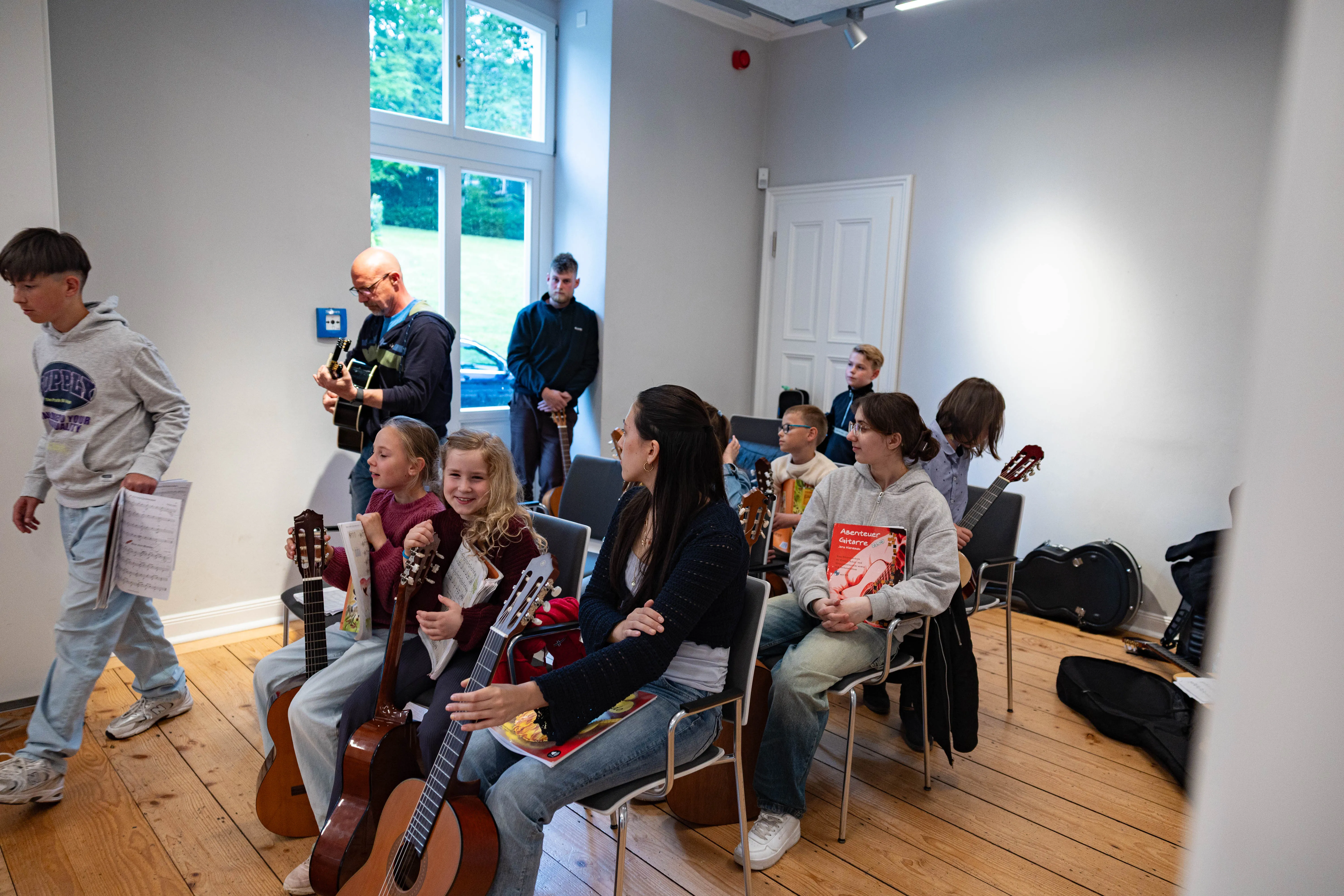 Mehrere Kinder und Jugendliche sitzen oder stehen in einem hellen Raum mit Gitarren und Notenblättern, während eine Frau eine Gitarre spielt und ein Mann im Hintergrund ebenfalls eine Gitarre hält.