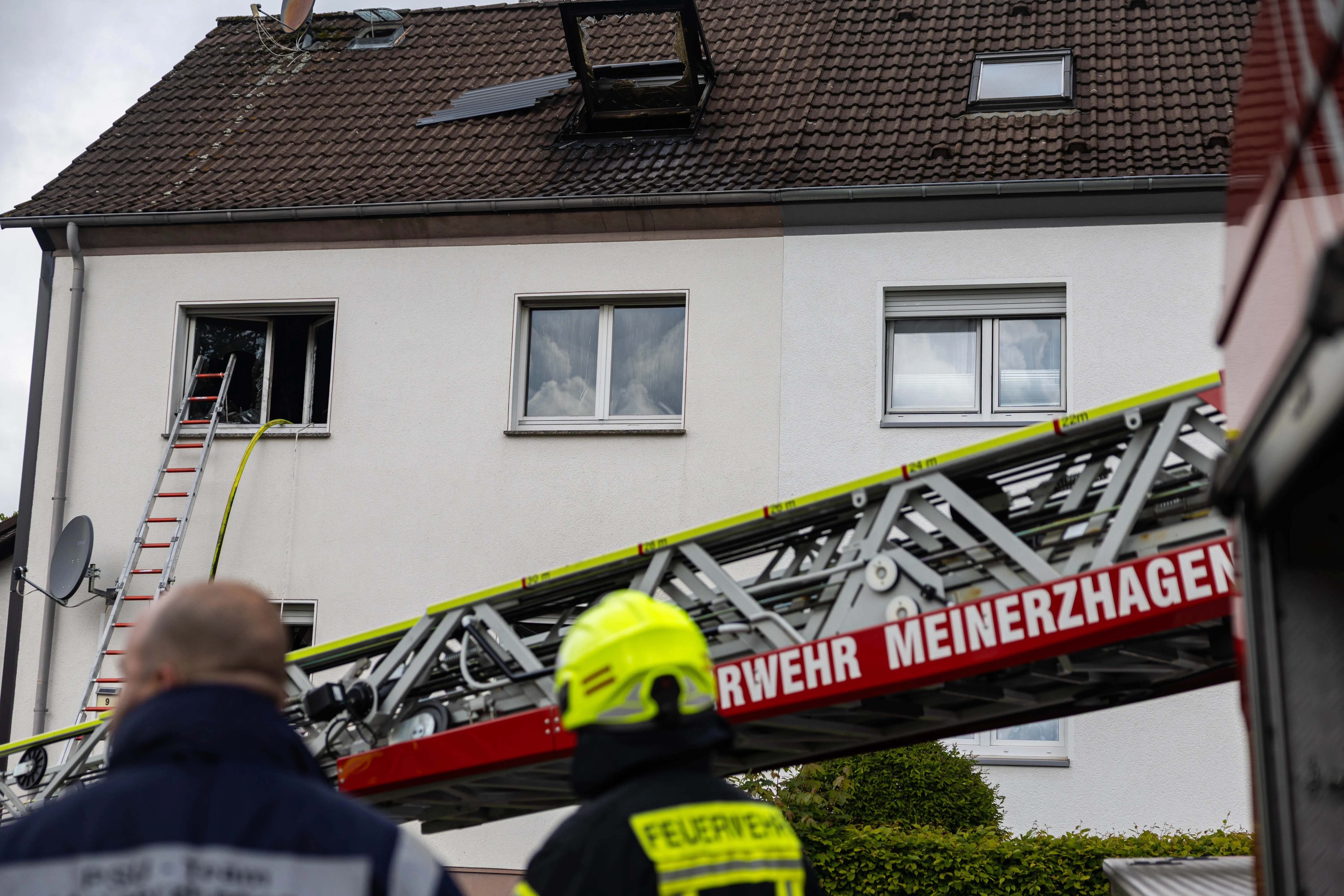 Feuerwehrleiterwagen der Feuerwehr Meinerzhagen steht vor einem zweigeschossigen Wohnhaus, dessen Fenster verrußt sind. Im Hintergrund sind Einsatzkräfte zu sehen.