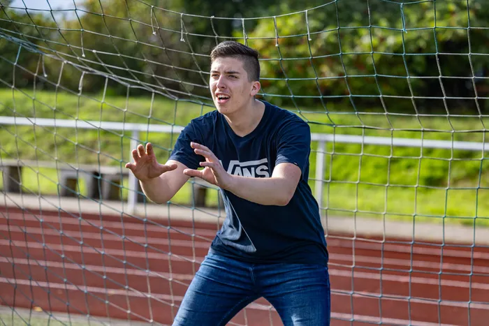 Ein junger Mann in einem dunkelblauen T-Shirt steht hinter einem Fußballnetz und hält die Hände in einer abwehrenden Geste bereit.