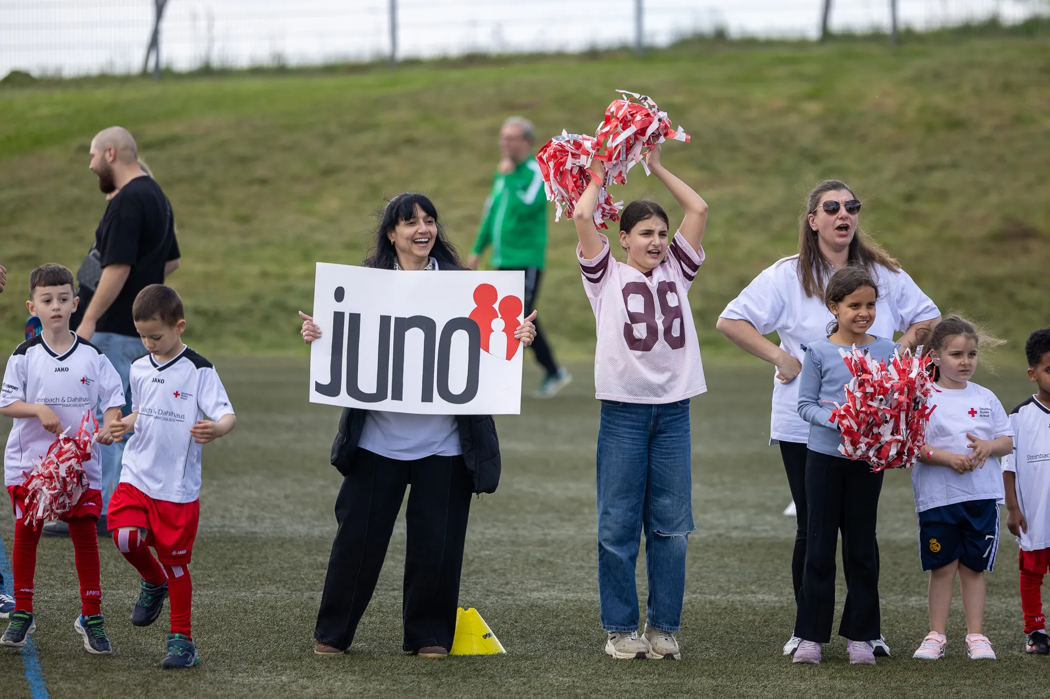 Zuschauer, darunter Kinder und Erwachsene, stehen an einem Fußballfeld und feuern an. Eine Frau hält ein Schild mit der Aufschrift „Juno“ hoch, ein Mädchen schwenkt Pompons.