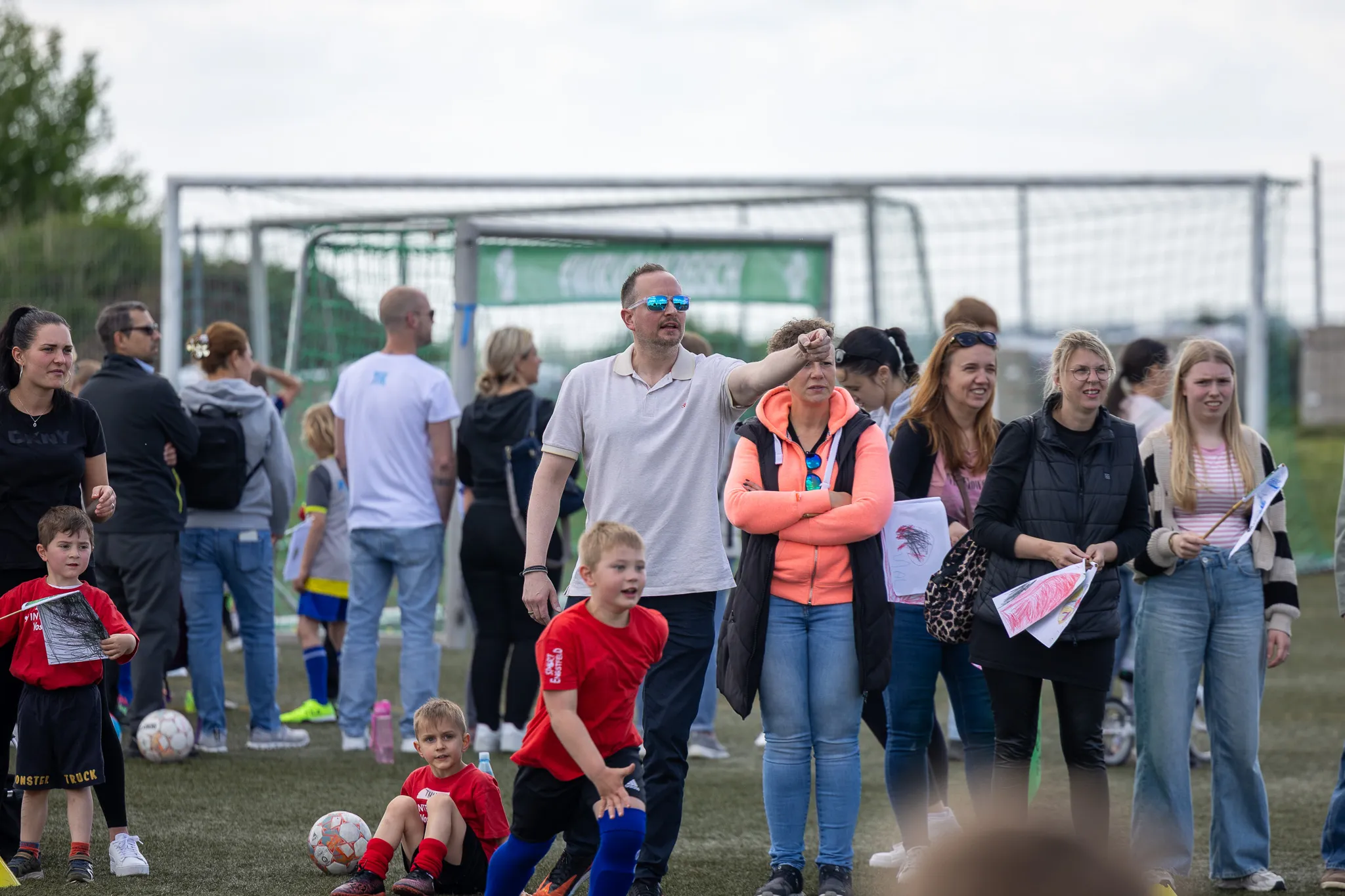 Zuschauer stehen und sitzen am Spielfeldrand eines Fußballturniers für Kinder, im Hintergrund ein Fußballtor.