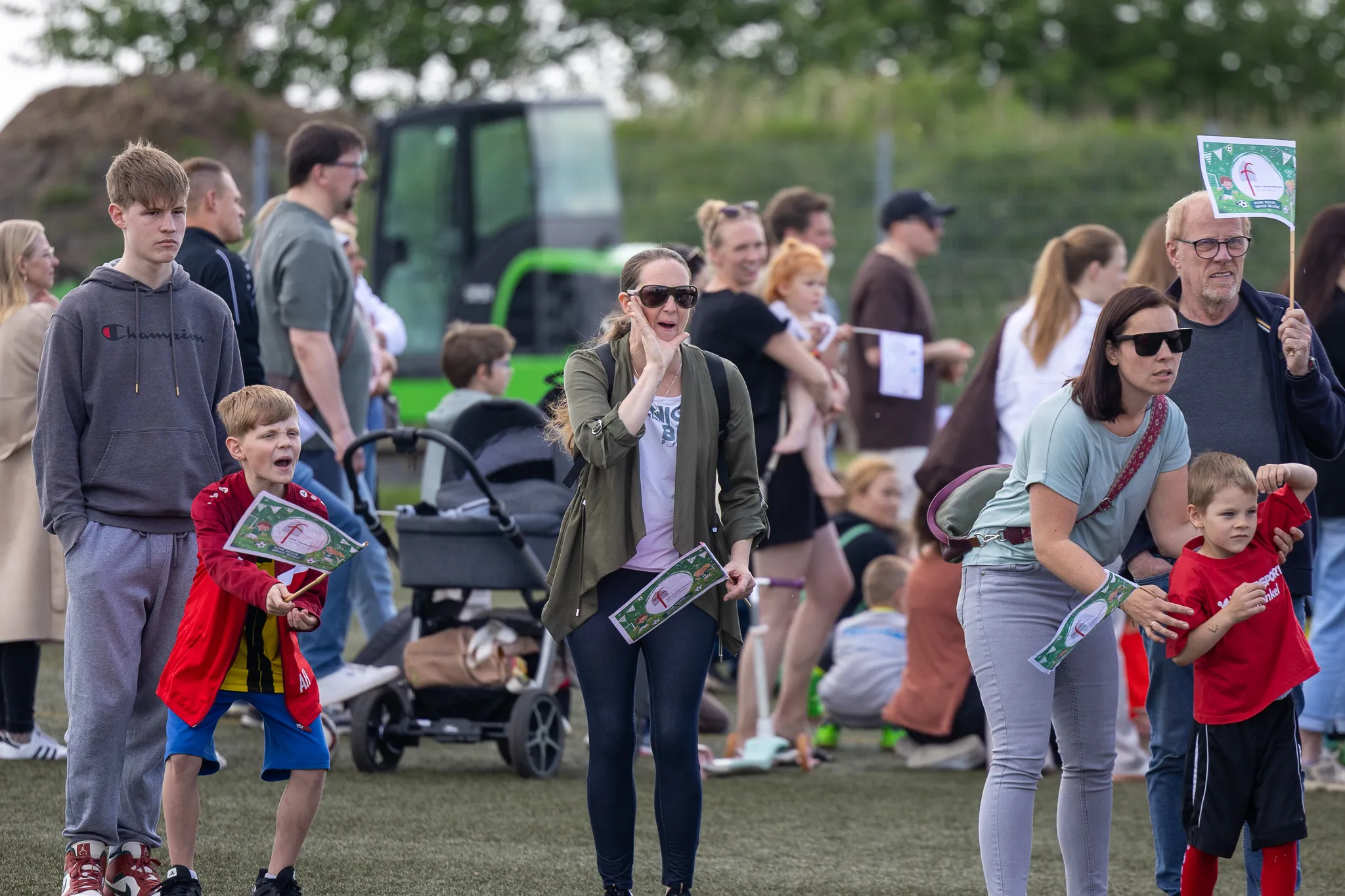 Zuschauer beobachten ein Fußballturnier für Kinder auf einer Wiese, im Hintergrund ein grüner Traktor.