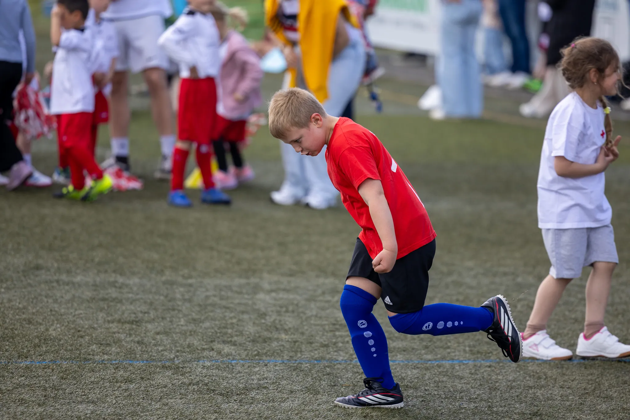Ein Junge in rotem Trikot und blauen Stutzen bereitet sich auf dem Rasen auf ein Fußballspiel vor, während andere Kinder im Hintergrund warten.