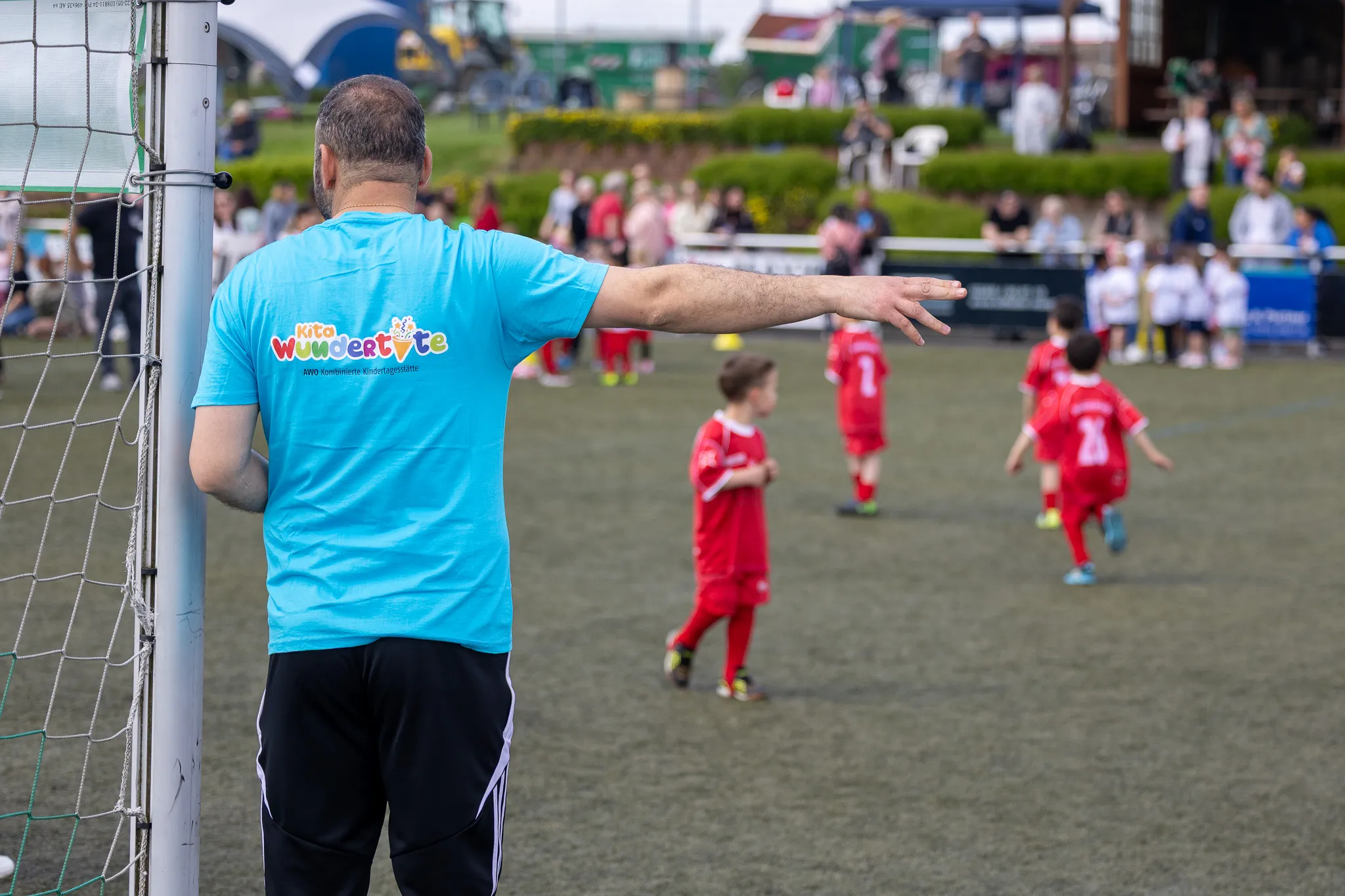 Ein Mann mit hellblauem T-Shirt und der Aufschrift „Kita Wundertüte“ deutet während eines Fußballspiels auf den Rasen. Im Hintergrund spielen Kinder in roten Trikots.