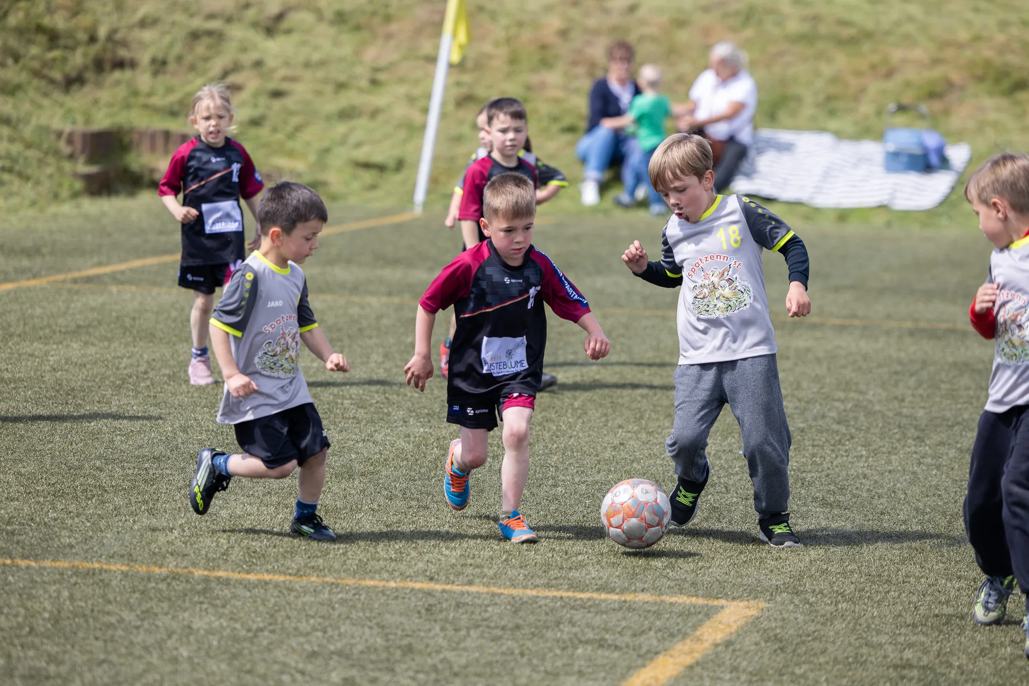 Mehrere Kinder spielen Fußball auf einem Rasenfeld, während Zuschauer im Hintergrund zusehen.
